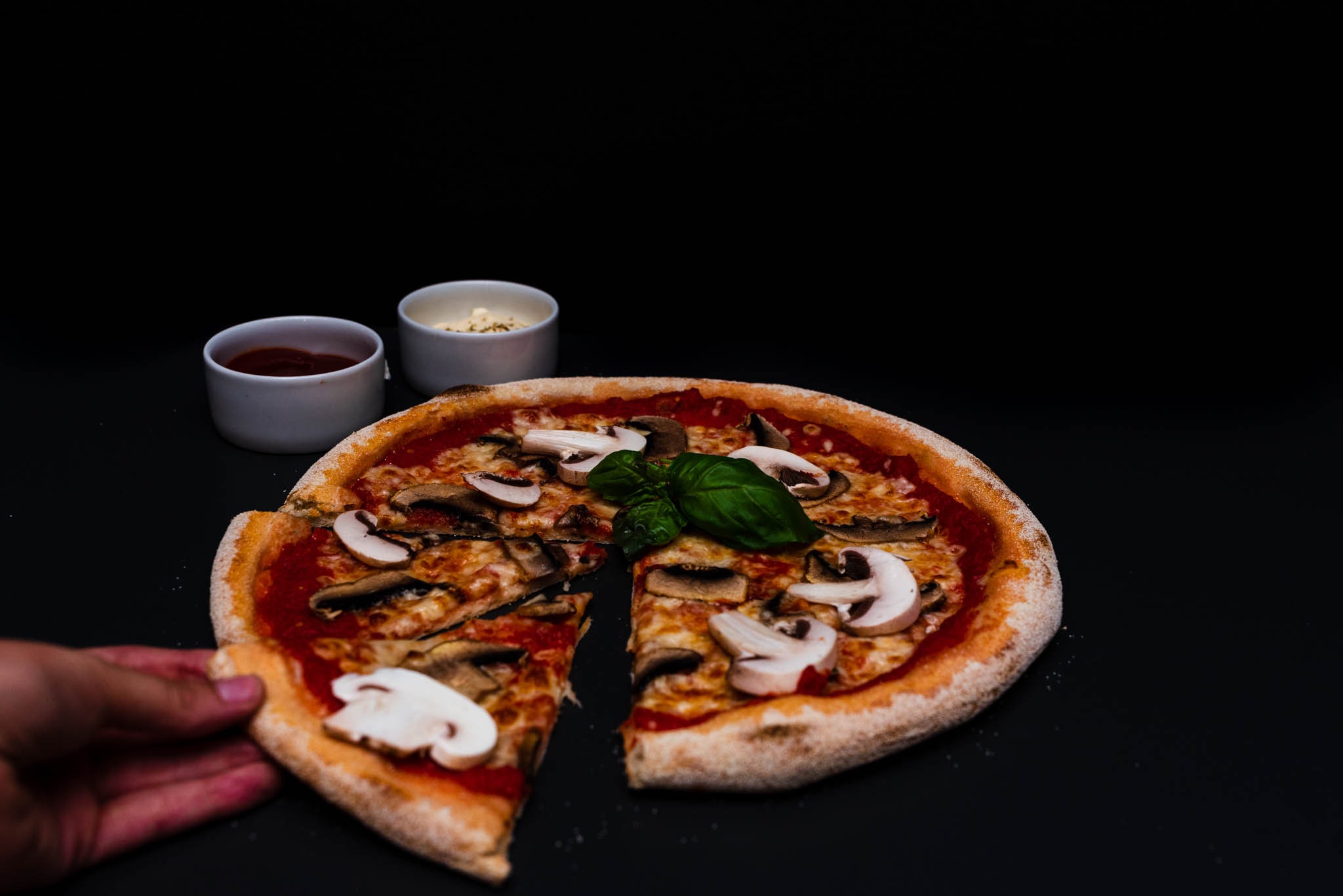 Pizza z pieczarkami na ciemnym tle, z dwoma sosami w białych miseczkach, prezentowana w Kluszkowcach. Kawałek pizzy jest podnoszony, ukazując spód ciasta.