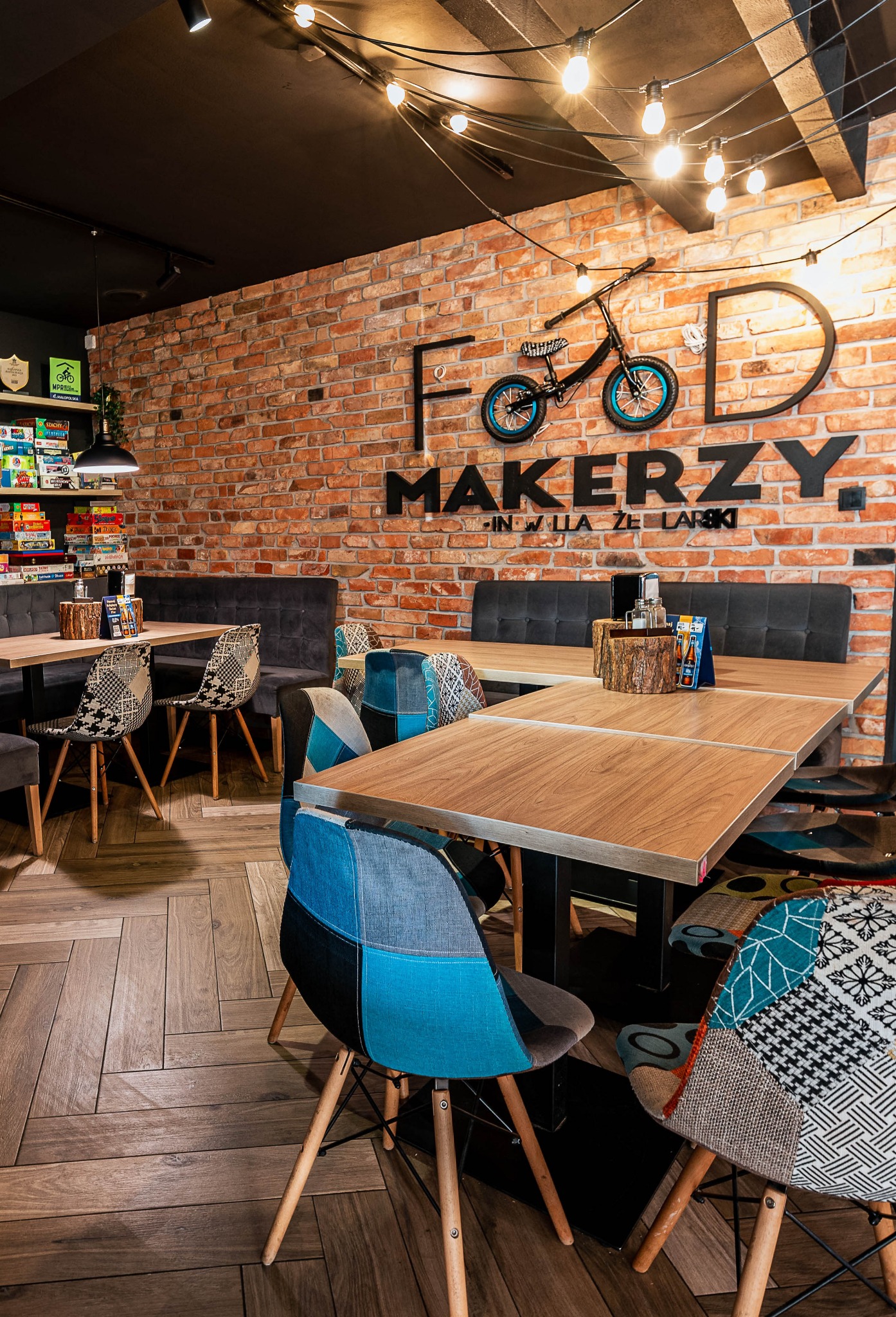 Wnętrze restauracji z ceglaną ścianą, drewnianymi stołami i krzesłami o patchworkowych wzorach, oświetlone dekoracyjnymi żarówkami i z logo 'Food Makerzy' z rowerem na ścianie.