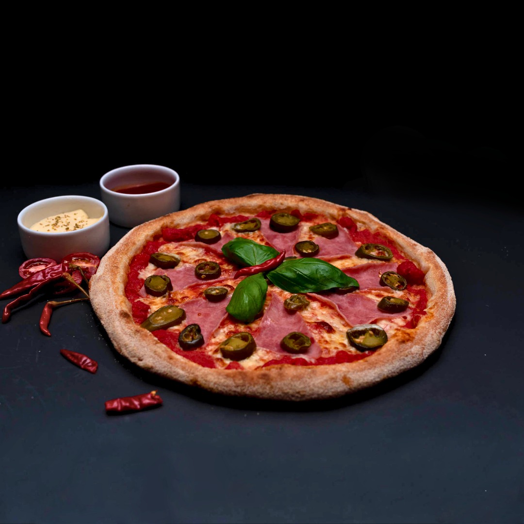 Pizza z szynką, papryczkami jalapeno i bazylią, podana z dwoma sosami na czarnym tle, udekorowana suszonymi papryczkami chili.