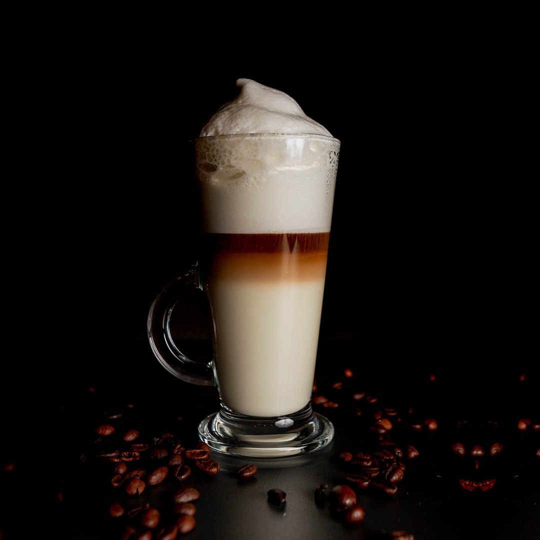 Wysoka szklanka z warstwową kawą latte zwieńczoną gęstą pianką, otoczona rozsypanymi ziarnami kawy na czarnym tle.