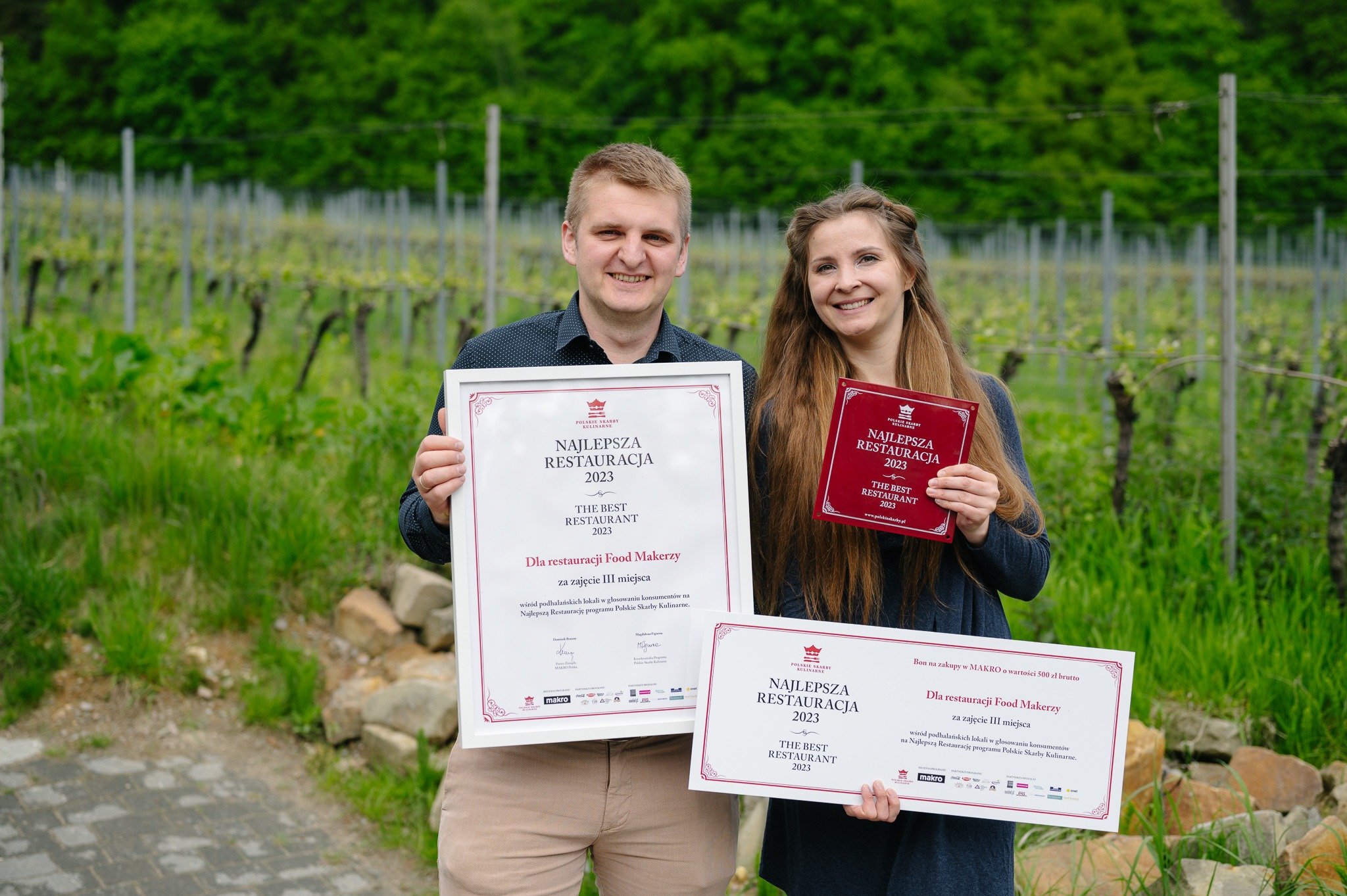 Dwie osoby prezentują nagrody dla restauracji Food Makerzy: dyplom w ramce, czerwoną tabliczkę i bon na zakupy, na tle winnicy.