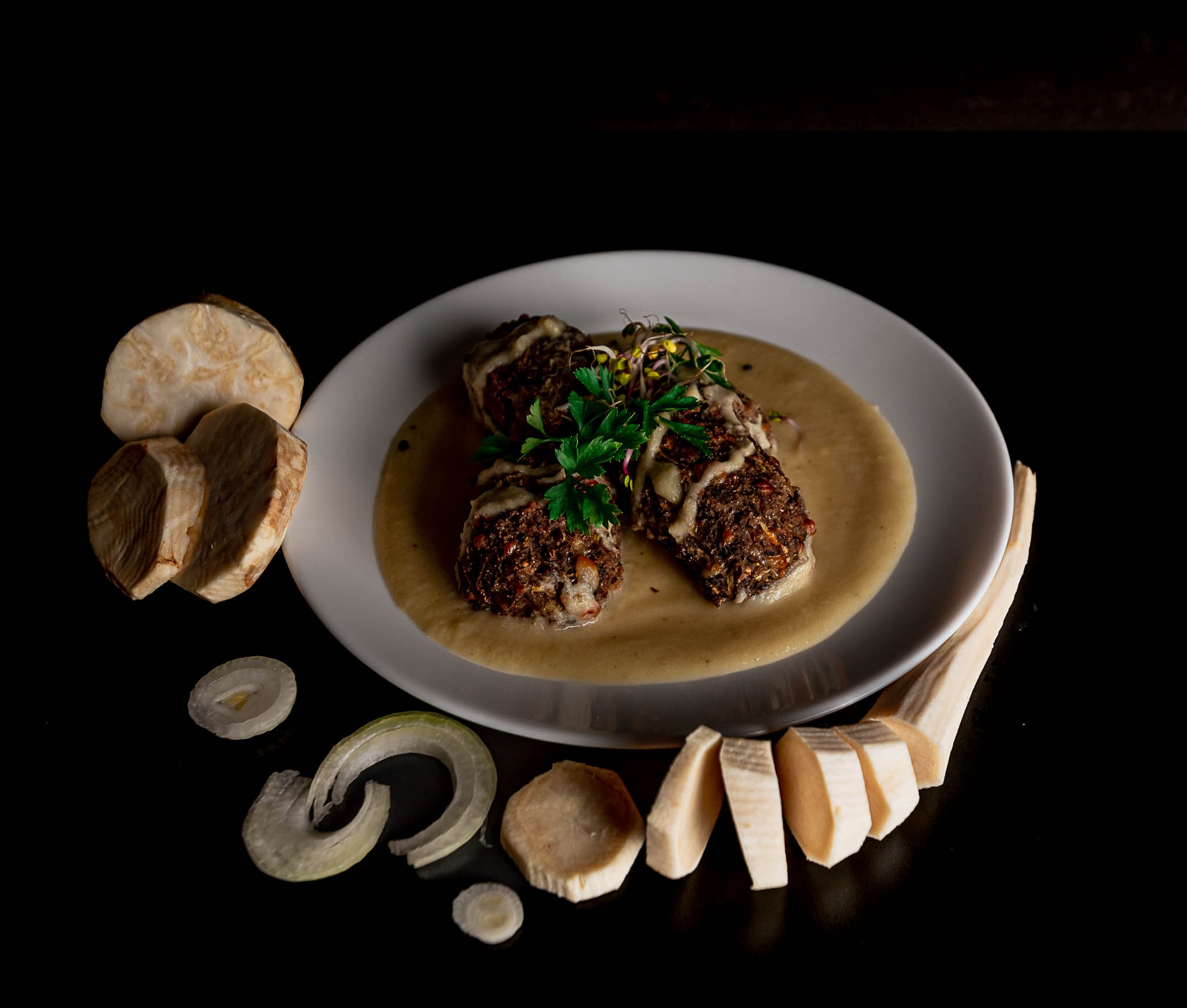 Wykwintne danie wegetariańskie: kotleciki w jasnym sosie, udekorowane świeżymi ziołami i kiełkami, podane na białym talerzu z plasterkami selera i cebuli na czarnym tle.