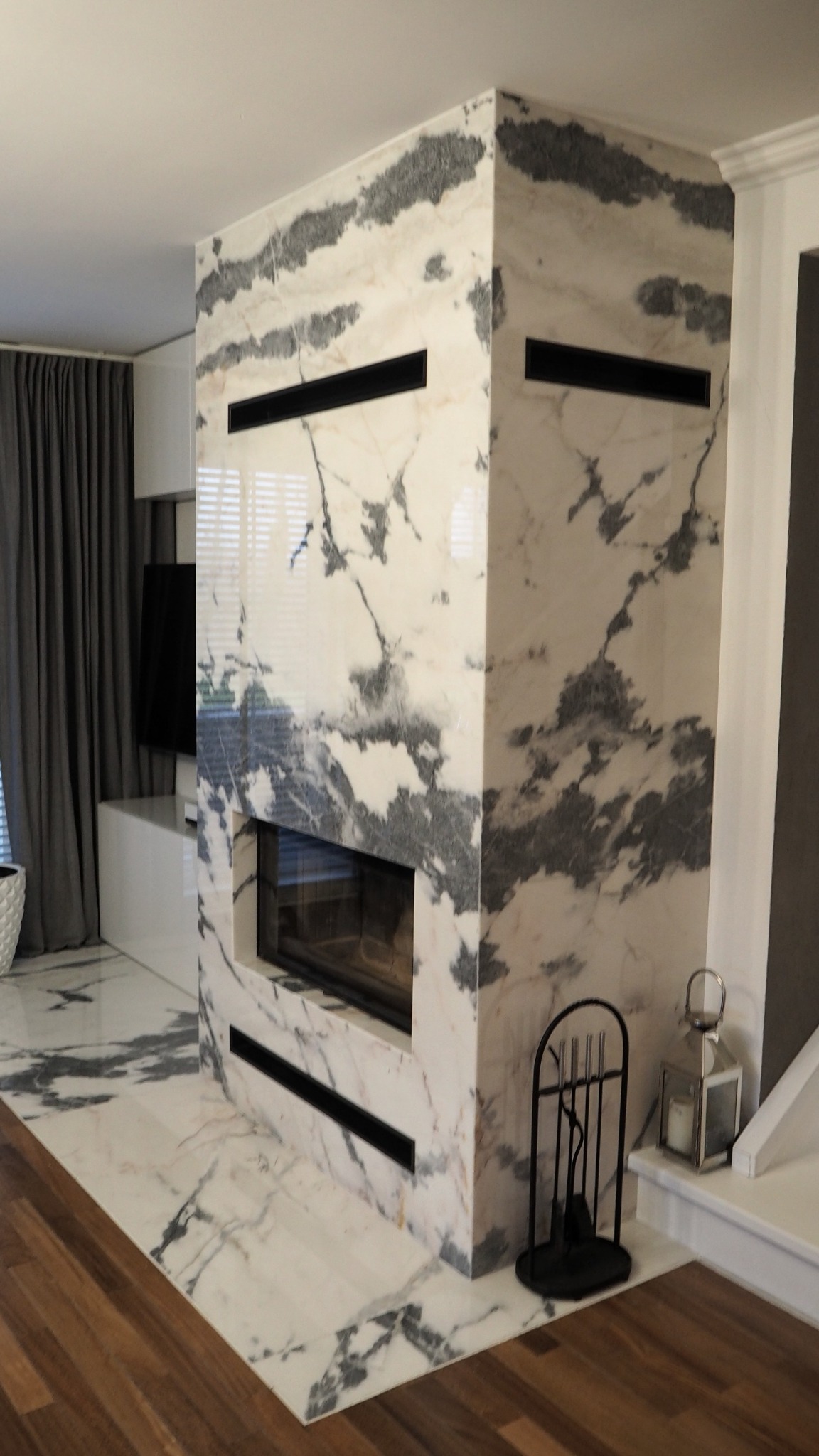 Nowoczesny kominek obłożony białym marmurem z szarymi żyłami, z czarnymi elementami wykończeniowymi, na podłodze z marmuru pasującego do kominka, z widocznymi akcesoriami kominkowymi.