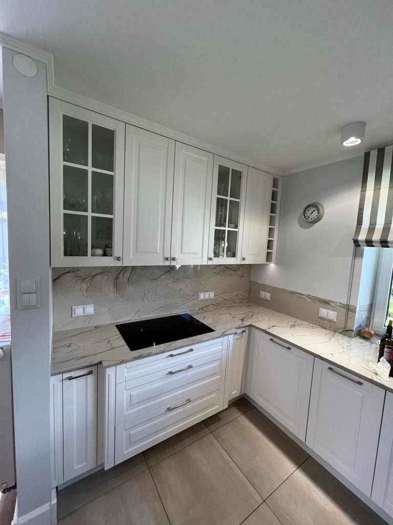 Jasna kuchnia z białymi szafkami i blatem imitującym marmur, płyta indukcyjna wkomponowana w blat, widok z lekkiego ukosa.