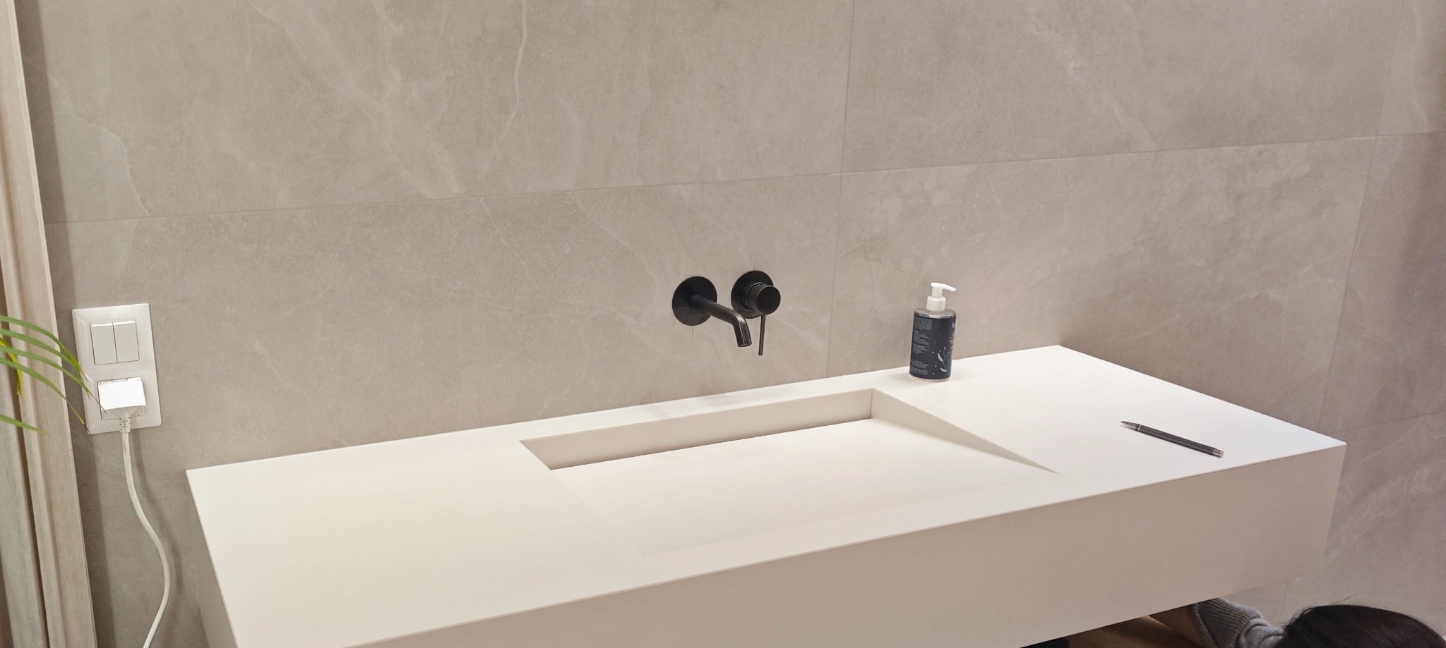 Biała, minimalistyczna umywalka zintegrowana z blatem w nowoczesnej łazience, z czarną armaturą i dozownikiem mydła, na tle płytek imitujących marmur.