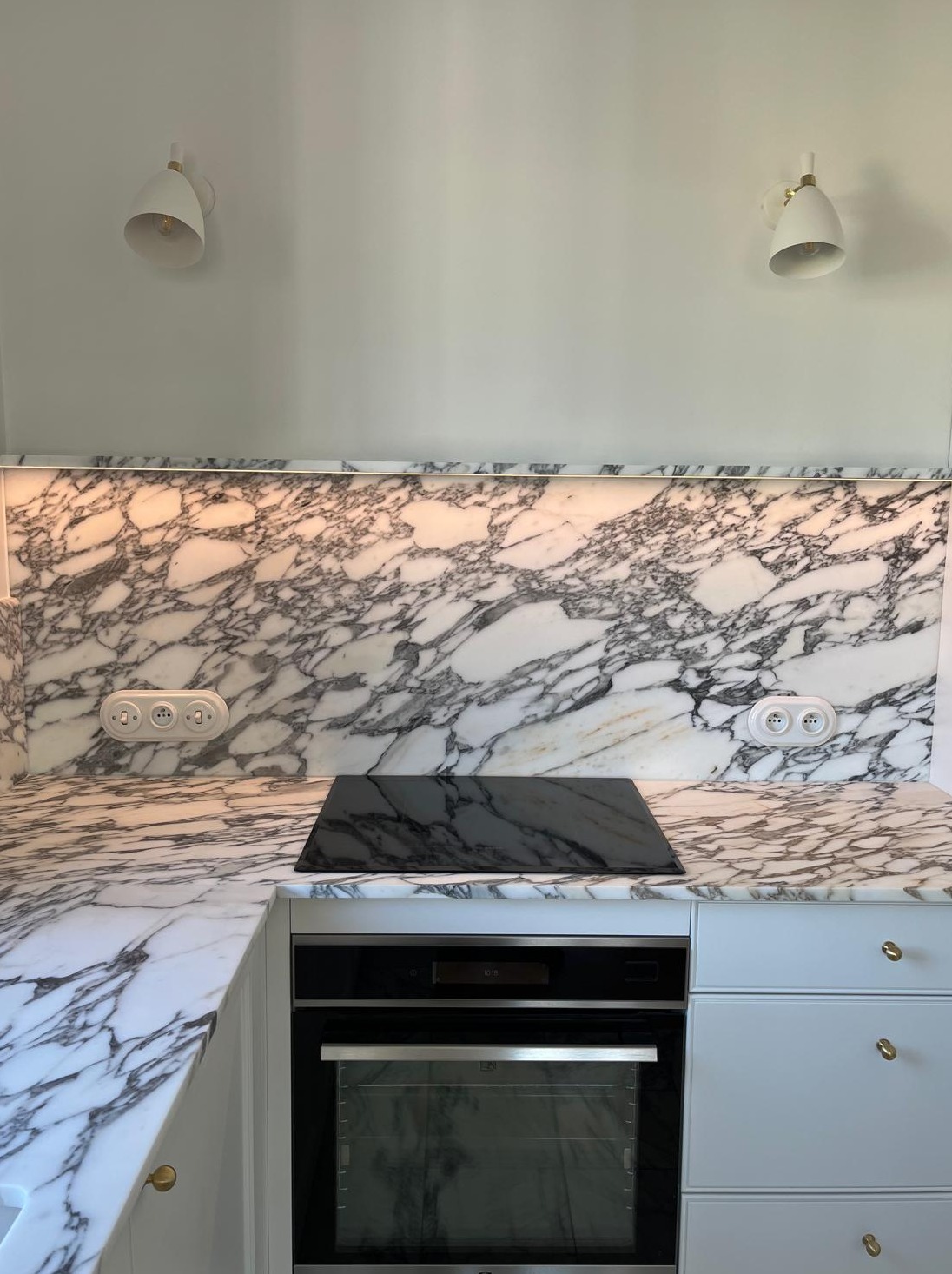 Kuchenny blat z marmuru Carrara z wbudowaną płytą indukcyjną i gniazdami elektrycznymi w stylu retro, oświetlony dwoma białymi lampami ściennymi.