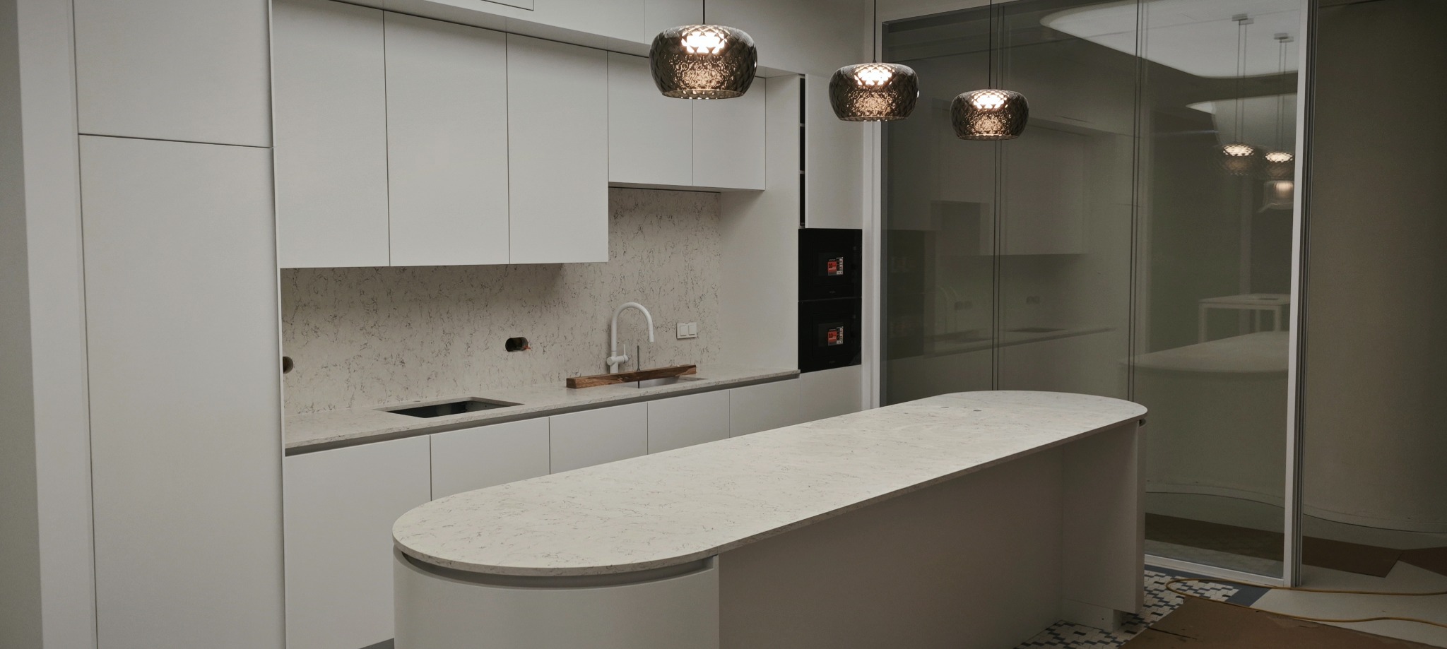Nowoczesna kuchnia w odcieniach bieli z wyspą i blatem o delikatnym wzorze, oświetlona trzema wiszącymi lampami o metalicznej powierzchni. Widoczny fragment instalacji wodnej nad blatem.