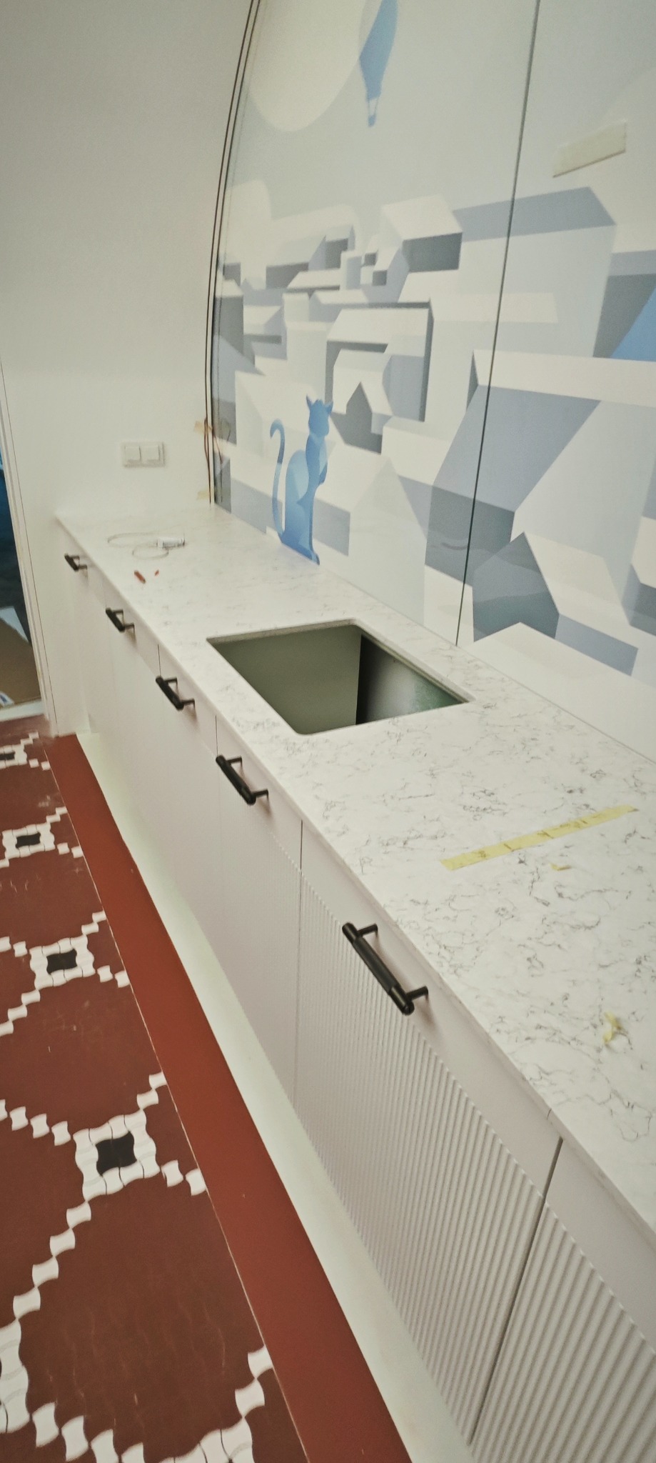 Nowoczesny biały blat z marmurowym wzorem, z otworem na zlew, zamontowany na białych szafkach z czarnymi uchwytami, na tle ściany z abstrakcyjnym rysunkiem miasta i niebieskiego kota