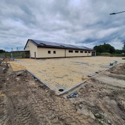 Mat-Bud - Parterowy budynek z panelami słonecznymi na dachu, widoczny z zewnątrz, z częściowo wyłożonym placem kostką. Niebo pochmurne, teren wokół w trakcie prac ziemnych.