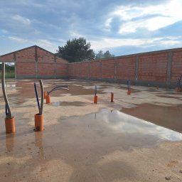 Mat-Bud - Wnętrze budynku w trakcie budowy z widocznymi ścianami z czerwonej cegły, instalacją wodną wystającą z mokrej posadzki i odbiciem nieba w kałużach.