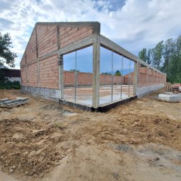 Mat-Bud - Surowy stan budynku z cegły i betonu w trakcie budowy, widoczne zbrojenia i fundamenty. Na pierwszym planie ziemia, w tle drzewa i pochmurne niebo.
