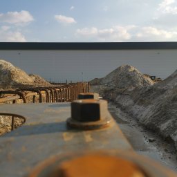 Mat-Bud - Z bliska: Zbrojenie fundamentów pod budynek przemysłowy. Widoczne stalowe pręty, śruby mocujące i wykop z piaskiem. W tle jasna elewacja hali i błękitne niebo.