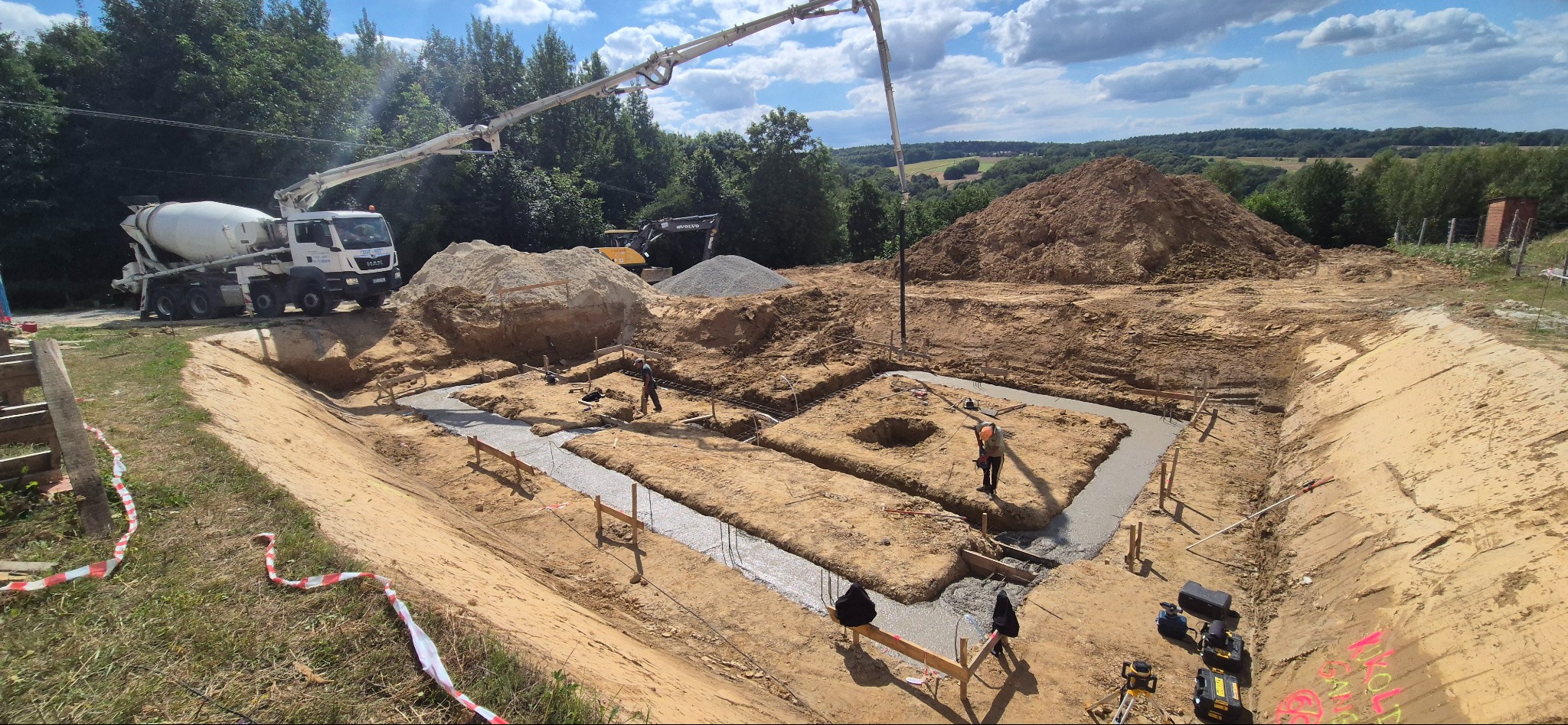 Wylewanie fundamentów domu jednorodzinnego z użyciem pompy do betonu. Widoczny wykop, szalunki i częściowo zalane ławy fundamentowe. W tle koparka i zieleń.