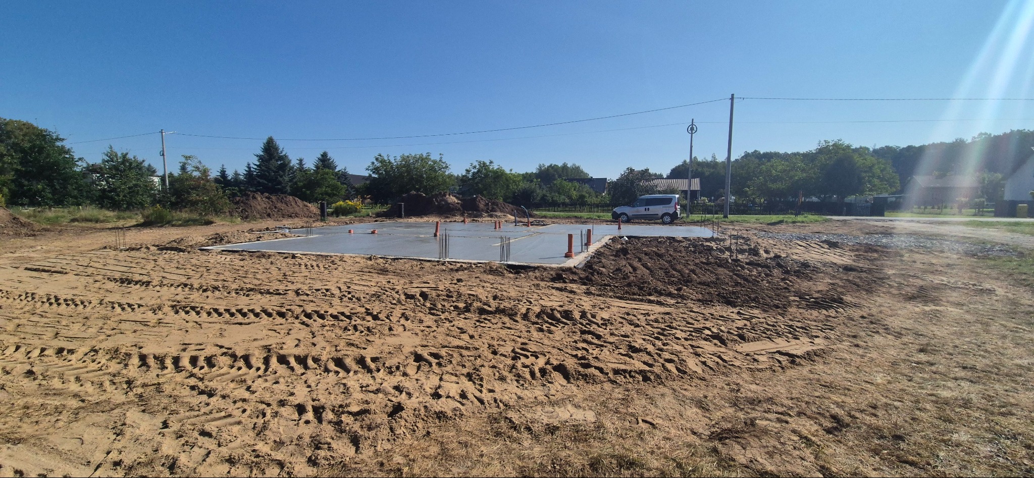 Wylana płyta fundamentowa z wystającymi prętami zbrojeniowymi i rurami w Kielanówce, na tle krajobrazu z samochodem i drzewami.