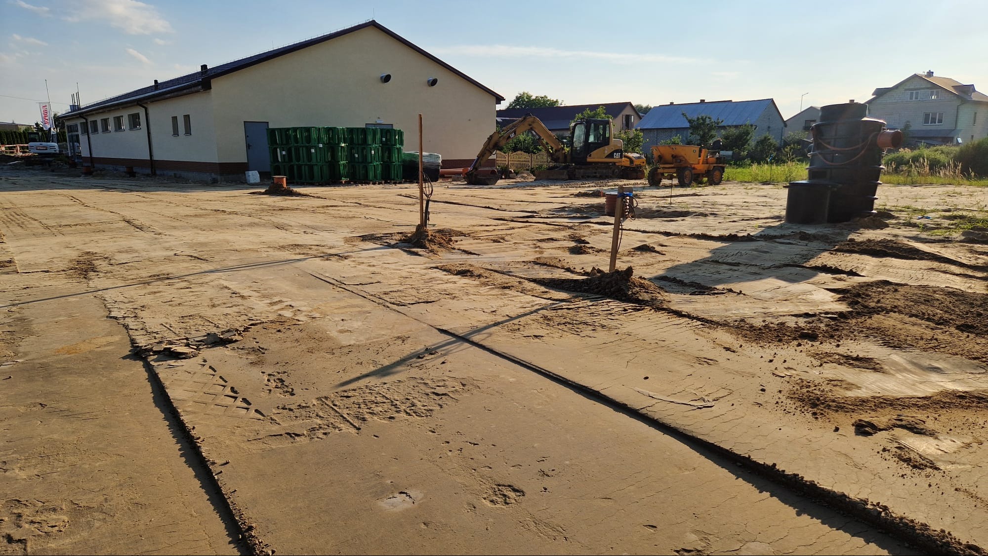 Przygotowanie terenu pod budowę: wyrównany teren z wykopami, maszyny budowlane w tle, zielone pojemniki i budynek gospodarczy w słoneczny dzień.