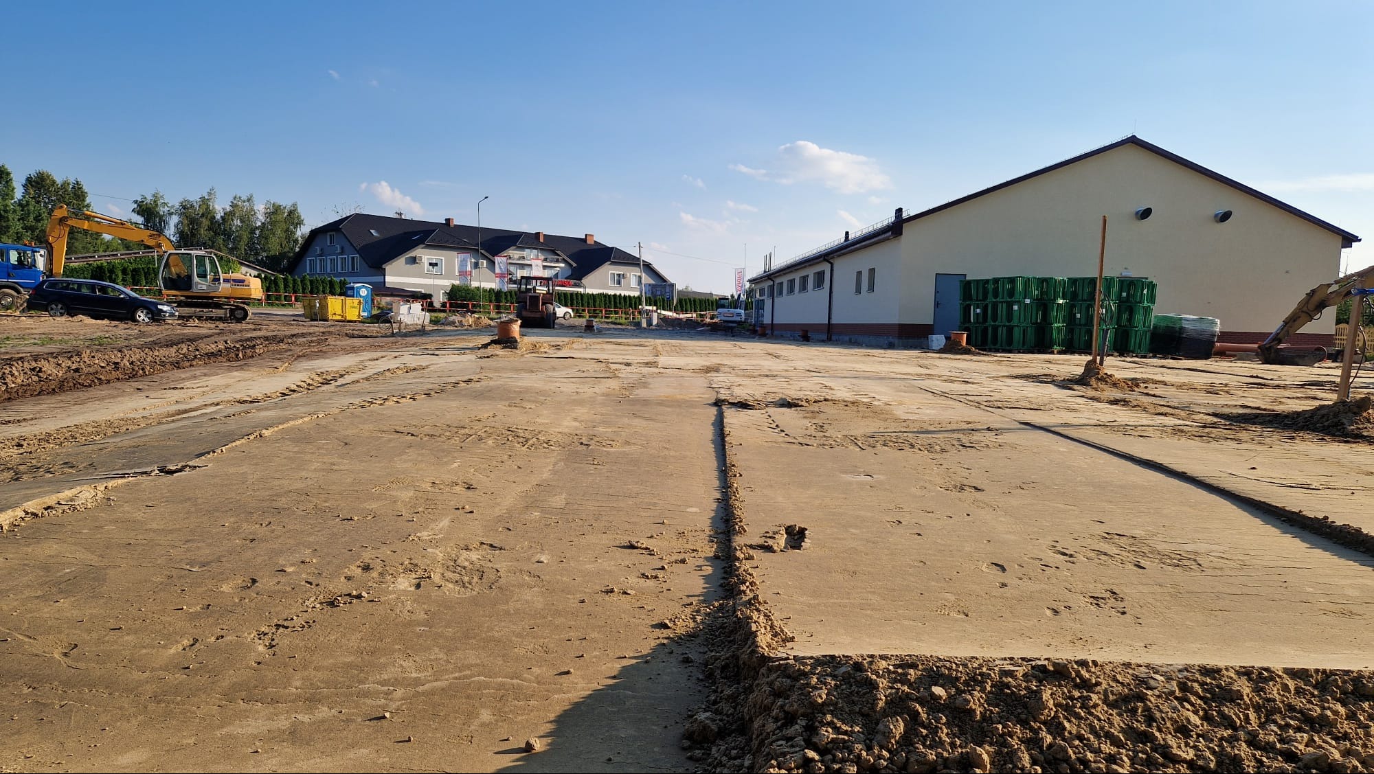 Przygotowanie terenu pod budowę: wykopane rowy, ciężki sprzęt (koparka, samochód) w tle, budynki w oddali, słoneczny dzień. Widok na plac budowy przed wylaniem fundamentów.