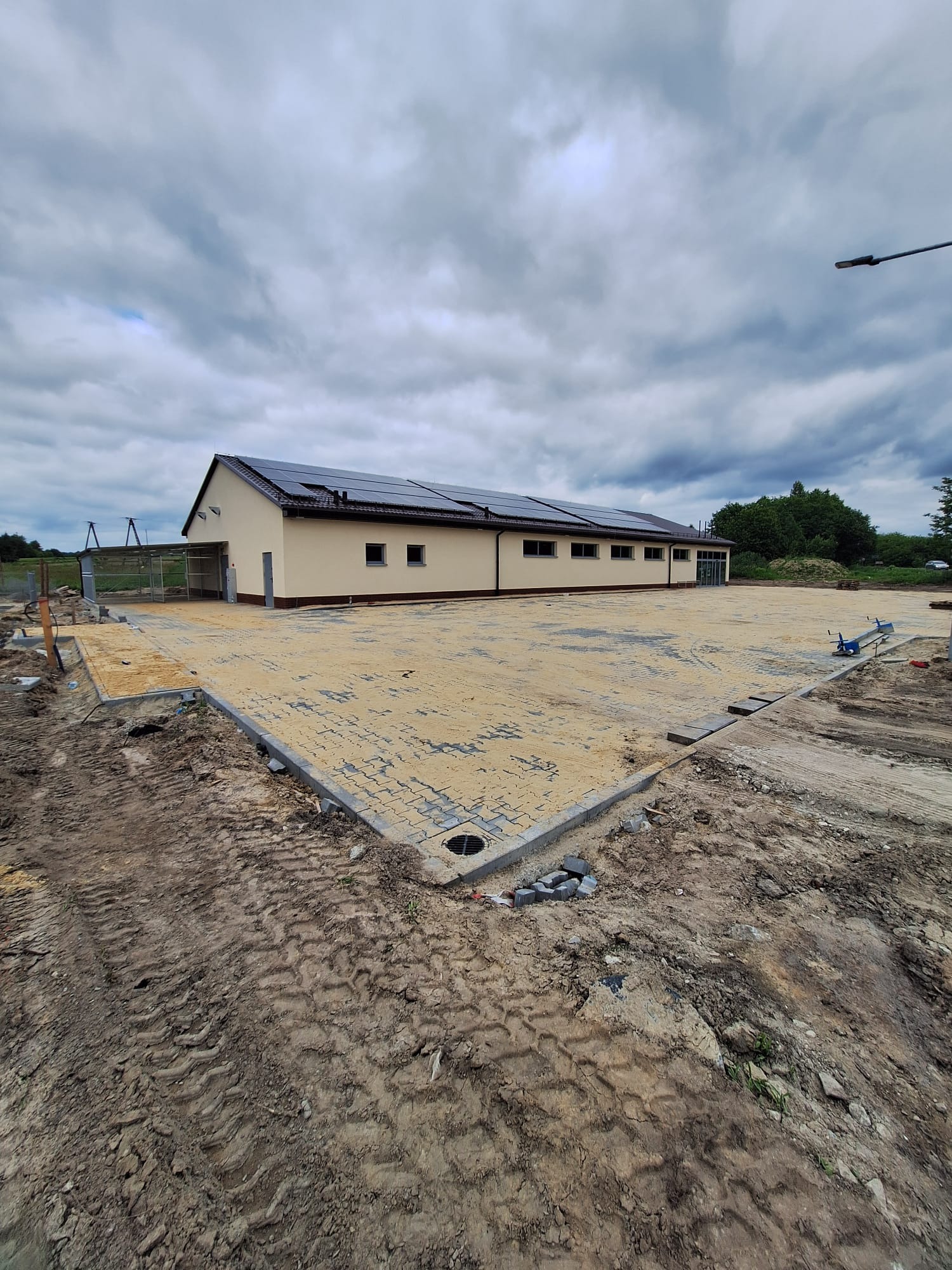 Parterowy budynek z panelami słonecznymi na dachu, widoczny z zewnątrz, z częściowo wyłożonym placem kostką. Niebo pochmurne, teren wokół w trakcie prac ziemnych.