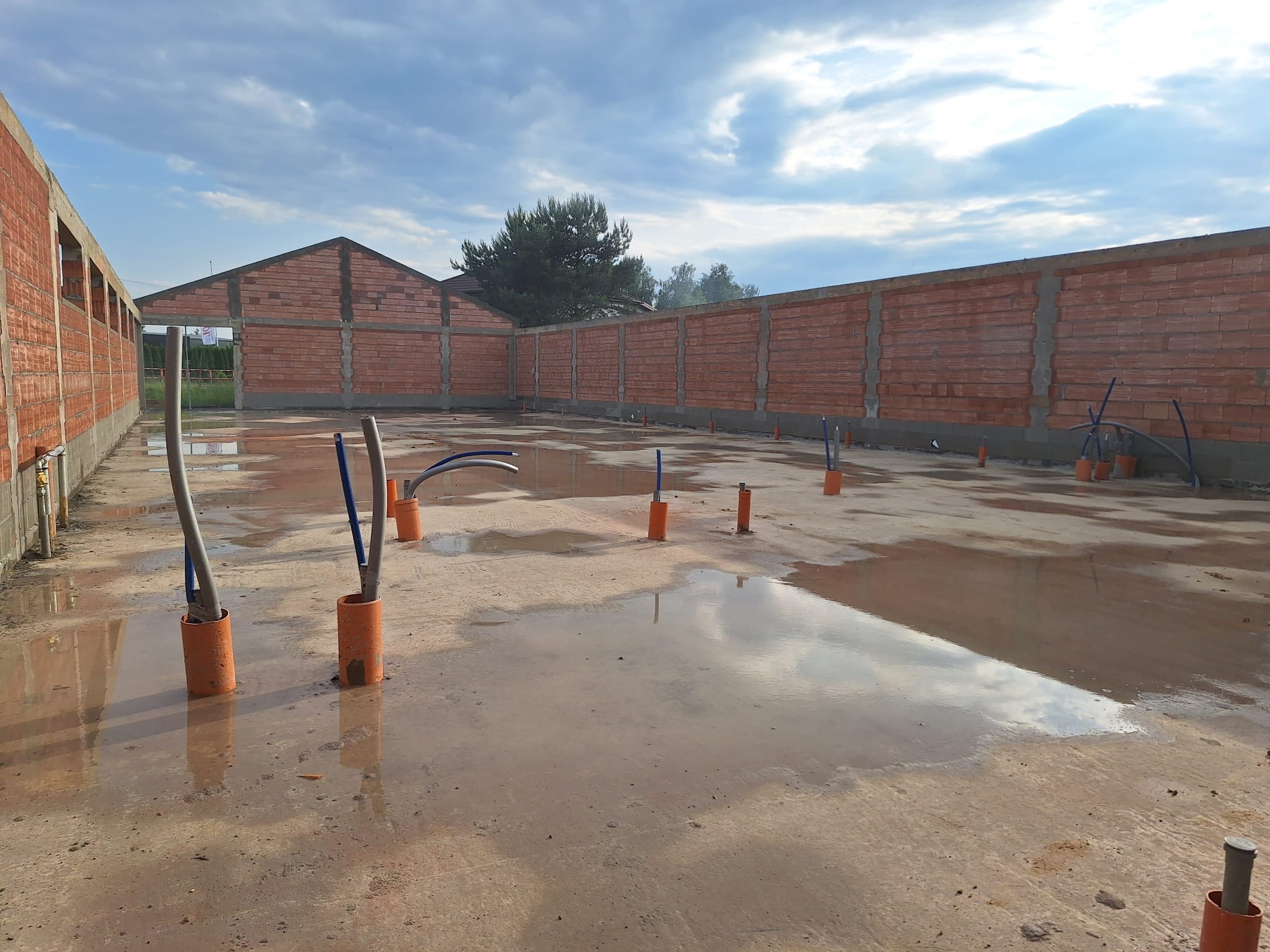 Wnętrze budynku w trakcie budowy z widocznymi ścianami z czerwonej cegły, instalacją wodną wystającą z mokrej posadzki i odbiciem nieba w kałużach.