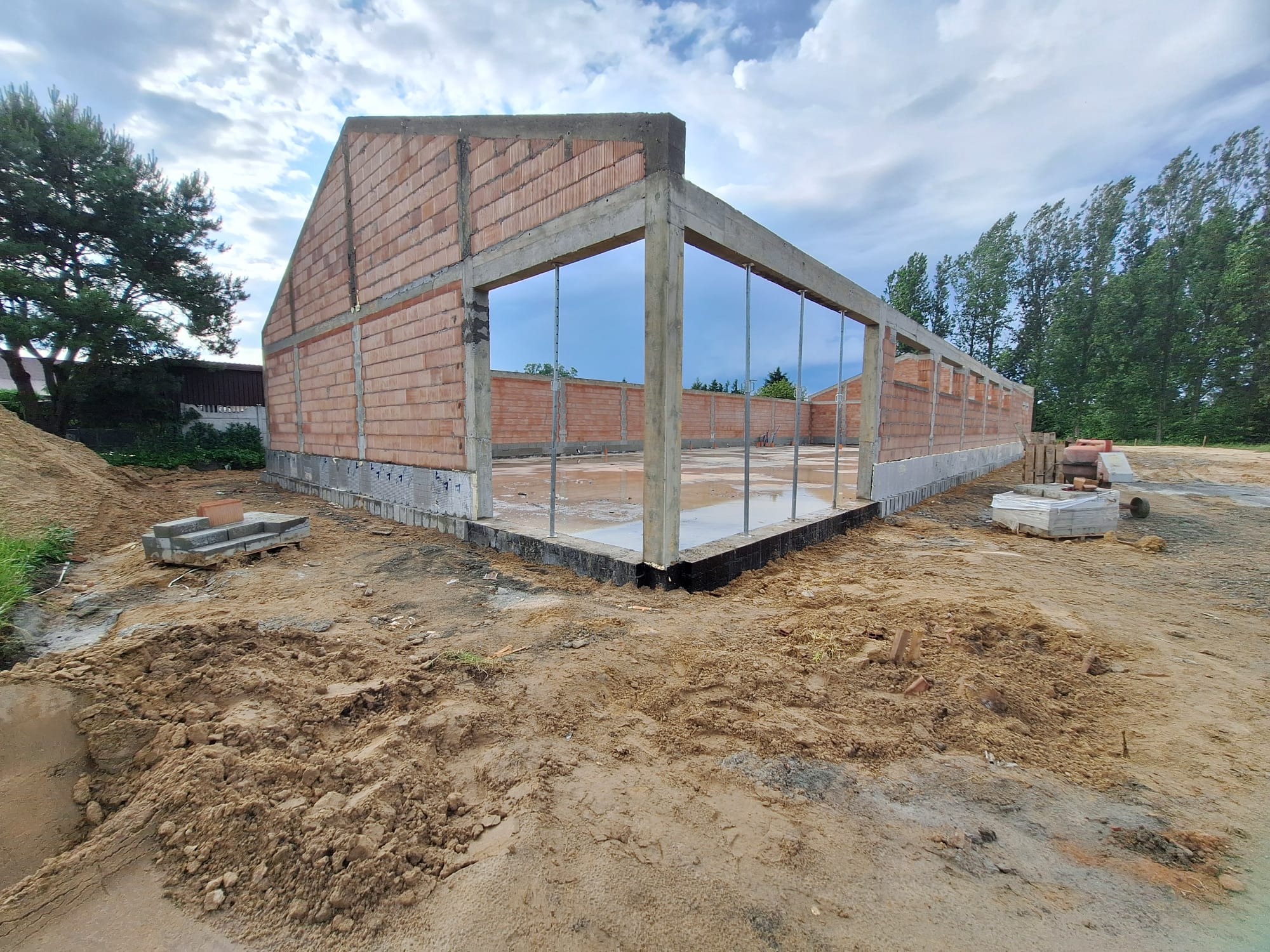 Surowy stan budynku z cegły i betonu w trakcie budowy, widoczne zbrojenia i fundamenty. Na pierwszym planie ziemia, w tle drzewa i pochmurne niebo.
