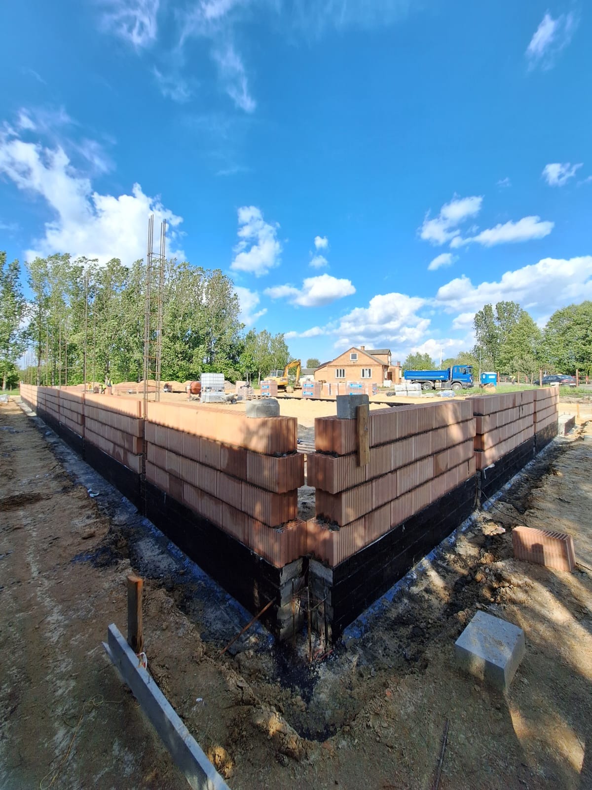 Wznoszenie ścian z czerwonej cegły na fundamencie z izolacją bitumiczną. Widoczne zbrojenie, w tle dom w budowie, błękitne niebo z chmurami i zieleń drzew.