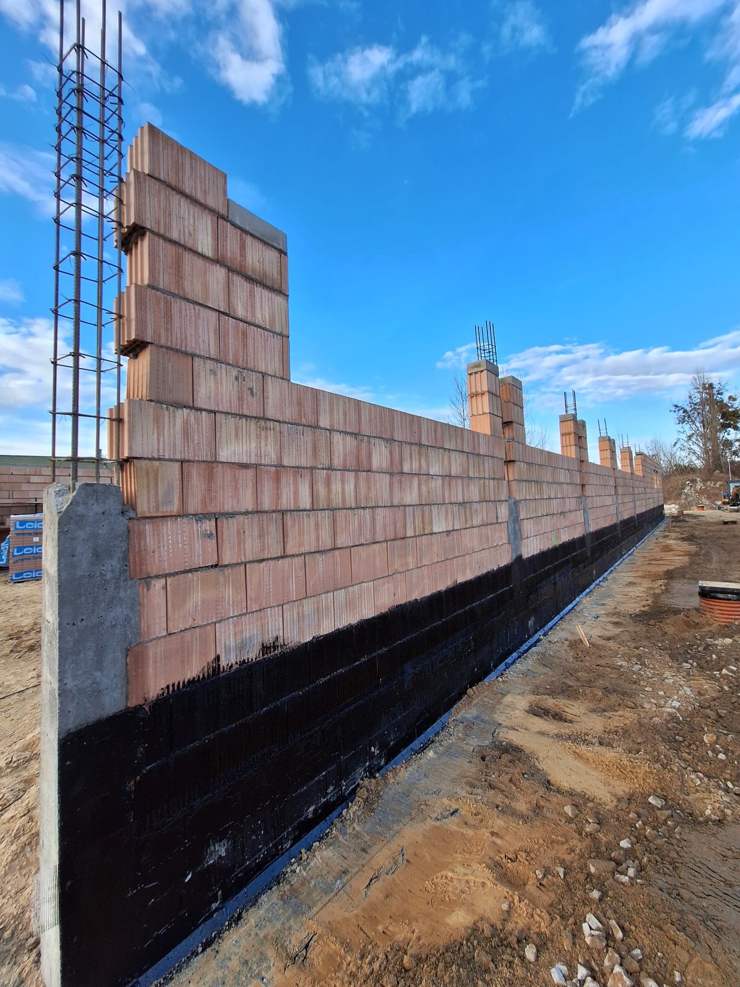 Wznoszenie ściany z pustaków ceramicznych z hydroizolacją fundamentów. Widoczne zbrojenie i niebo z chmurami. Prace murarskie w toku.