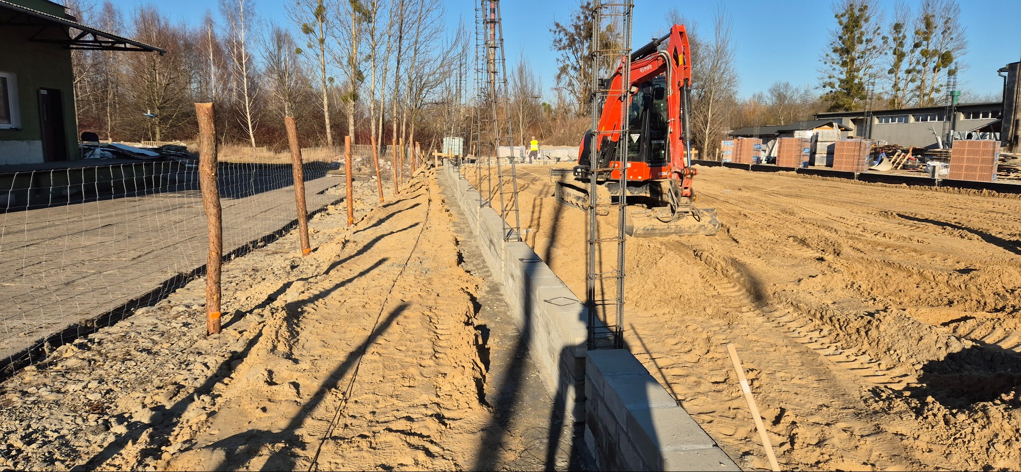 Fundamenty z bloczków betonowych i zbrojenia, wykop pod fundament, koparka na placu budowy, w tle palety z cegłą. Widok na budowę w słoneczny dzień.