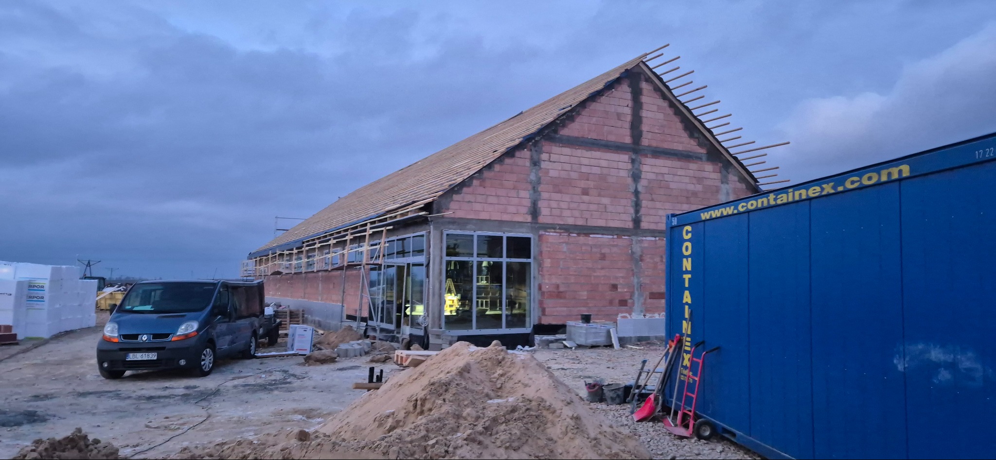 Budowa domu z czerwonej cegły w trakcie realizacji, widoczne okna, drewniana konstrukcja dachu, kontener i samochód dostawczy na placu budowy, pochmurne niebo.