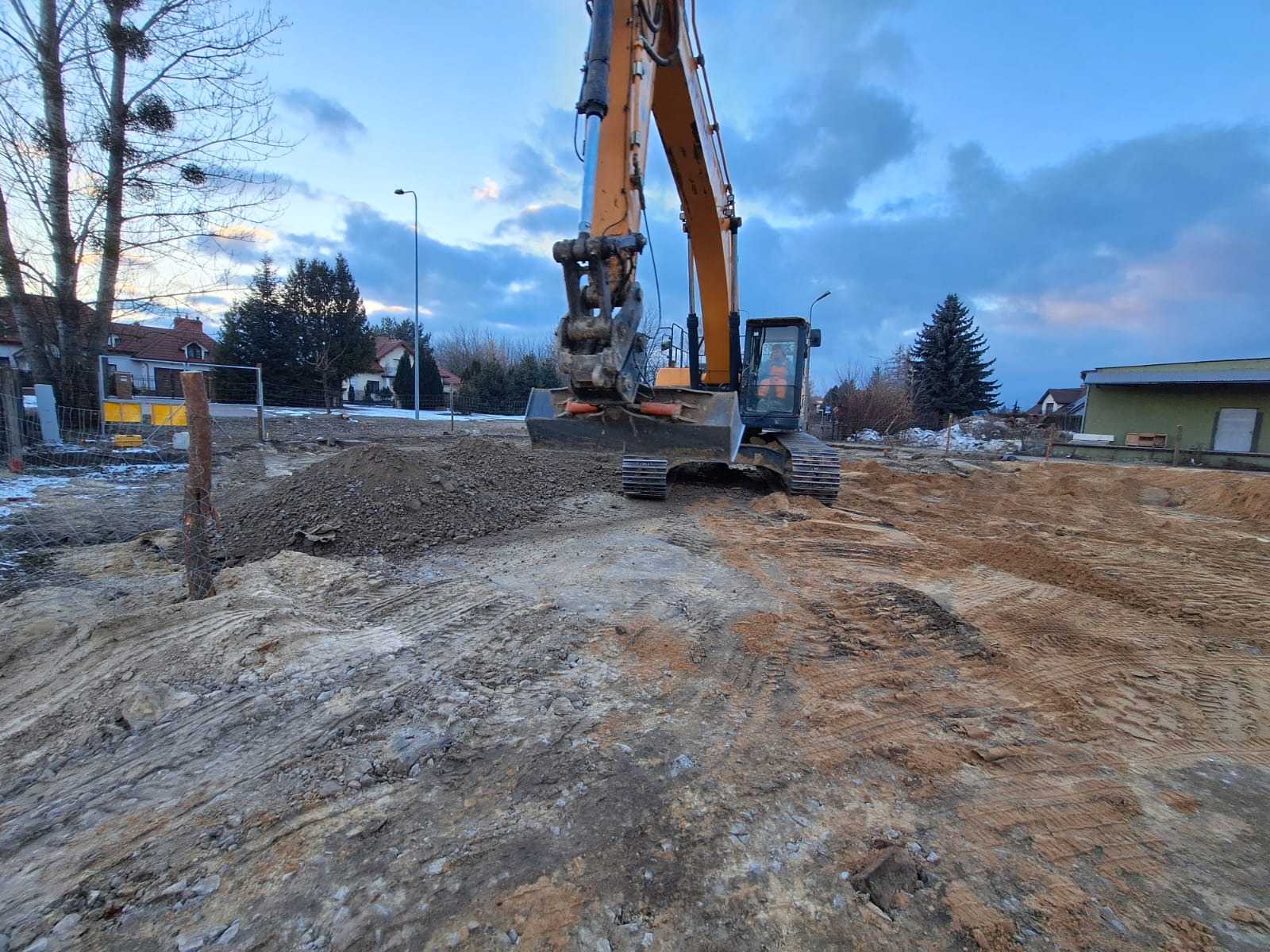 Żółta koparka na placu budowy z ziemią i piaskiem, w tle budynki mieszkalne i pochmurne niebo. Widoczne ślady opon na ziemi.
