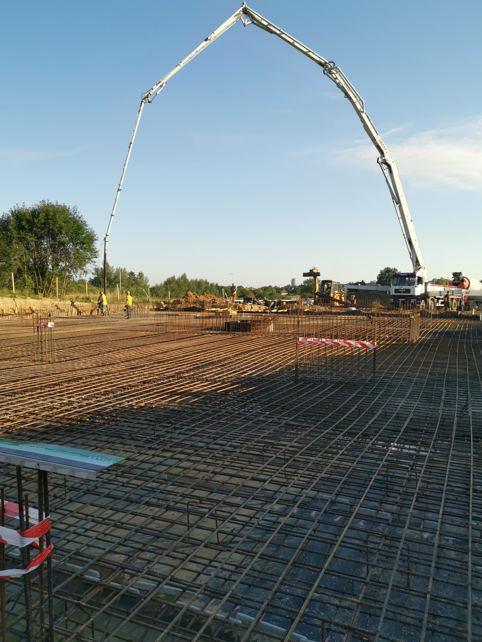 Zbrojenie fundamentów pod budowę, widoczna pompa do betonu i pracownicy w oddali. Siatka zbrojeniowa ułożona na przygotowanym terenie, w tle drzewa i błękitne niebo.