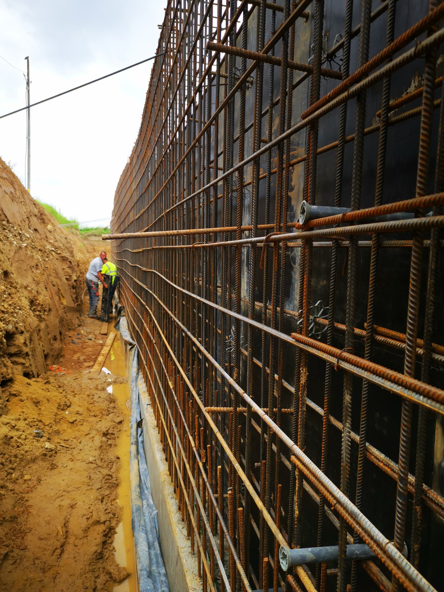 Zbrojenie pionowej ściany szalunkowej z widocznymi pracownikami w wykopie. Element konstrukcyjny w trakcie budowy, z bliska, z rdzawymi prętami i czarnym szalunkiem.