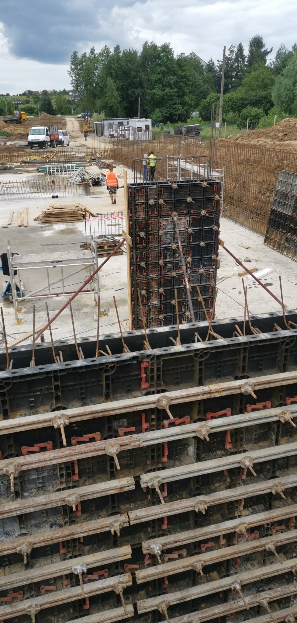 Szczegółowe ujęcie szalunków ścian zbrojonych na placu budowy, z widocznymi elementami konstrukcyjnymi i pracownikami w tle. Praca w toku, dzień pochmurny.