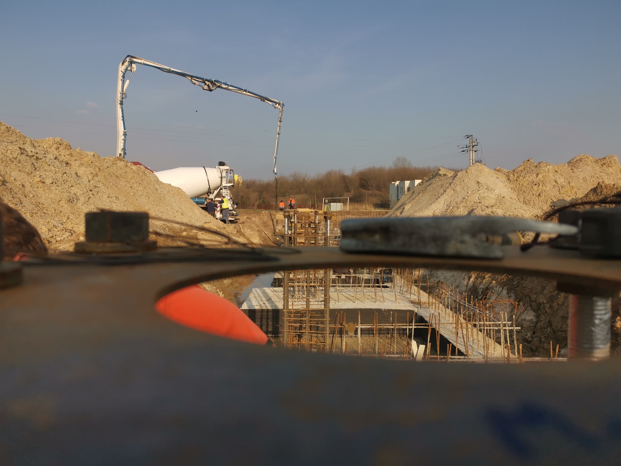 Betonowanie fundamentów pod budowę. Widok przez otwór w metalowej konstrukcji na zbrojenie i wylewany beton z betonomieszarki. Dwóch pracowników w odblaskowych ubraniach.