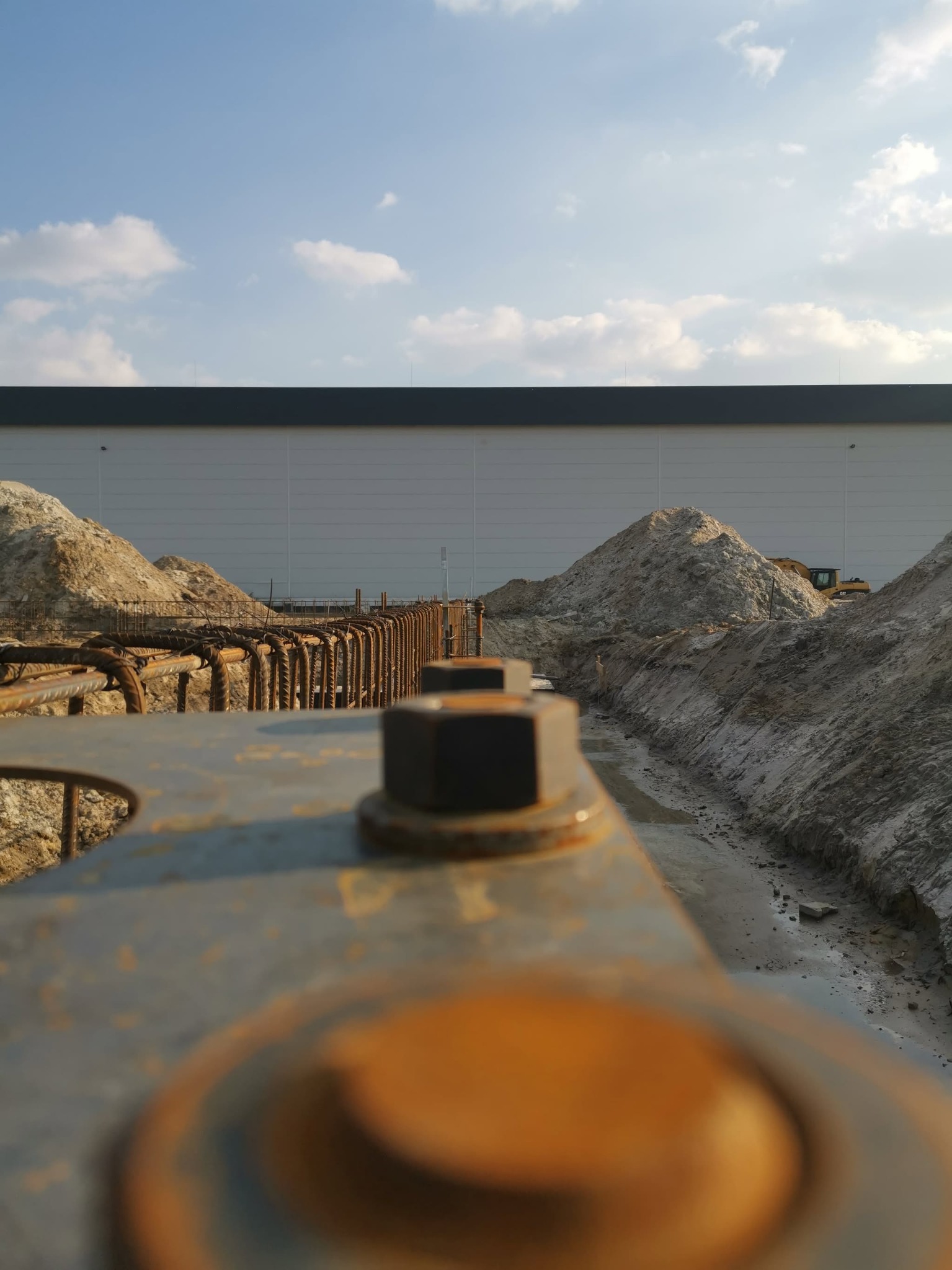 Z bliska: Zbrojenie fundamentów pod budynek przemysłowy. Widoczne stalowe pręty, śruby mocujące i wykop z piaskiem. W tle jasna elewacja hali i błękitne niebo.