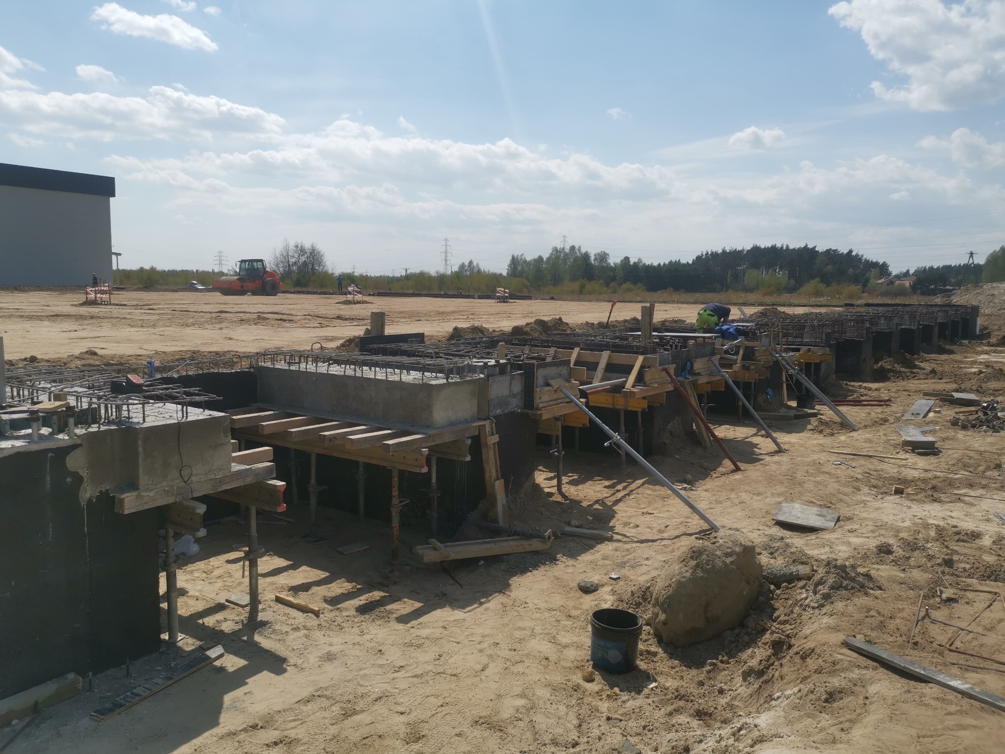 Wykopy pod fundamenty zbrojone stalą i deskowaniem, widok na plac budowy w Kielanówce w słoneczny dzień. W tle maszyna budowlana i budynek.