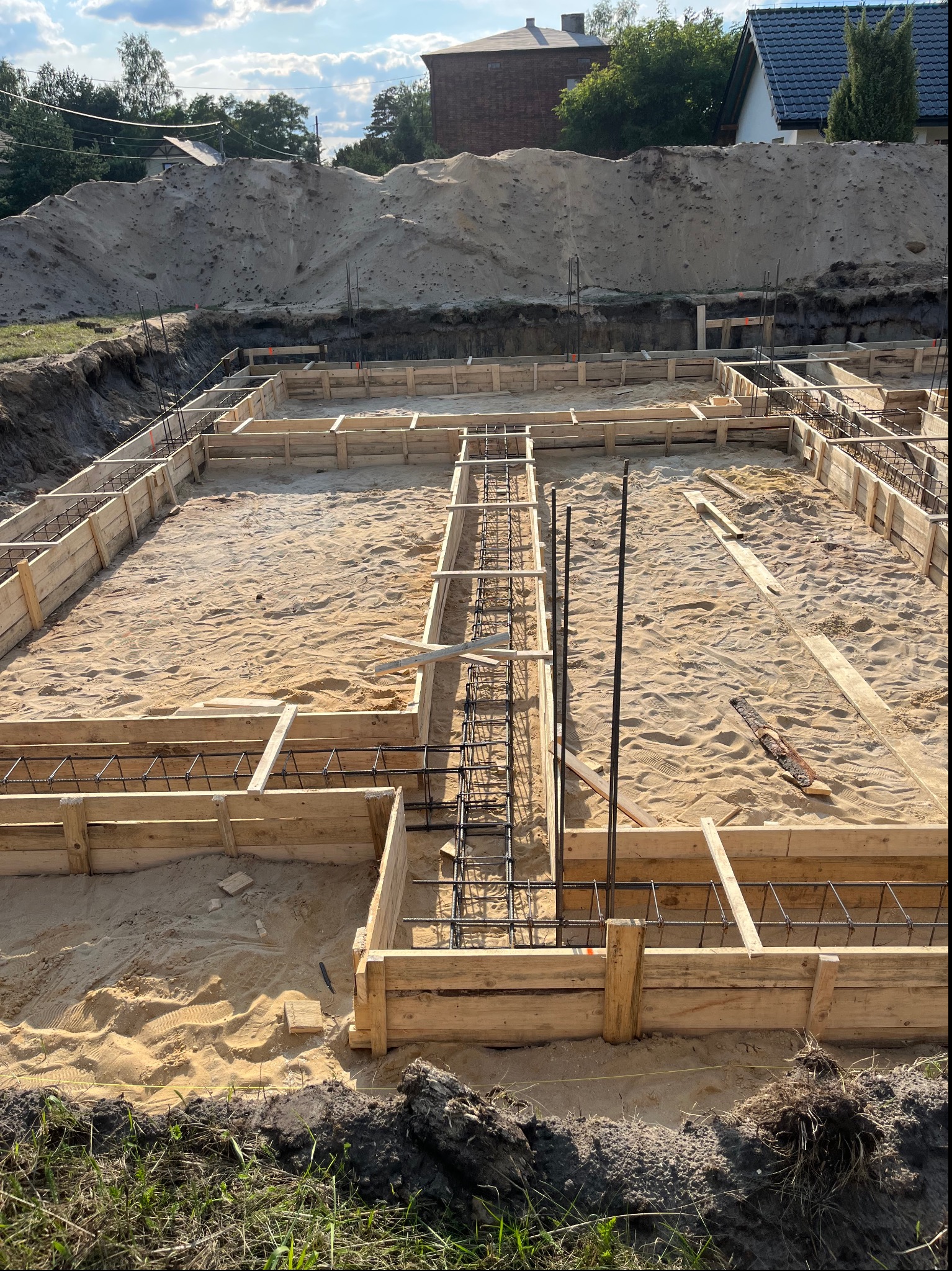 Przygotowanie terenu pod budowę domu: szalunek z desek i stalowe zbrojenie na piaszczystym podłożu, w tle pryzma piasku i zabudowa jednorodzinna pod błękitnym niebem.