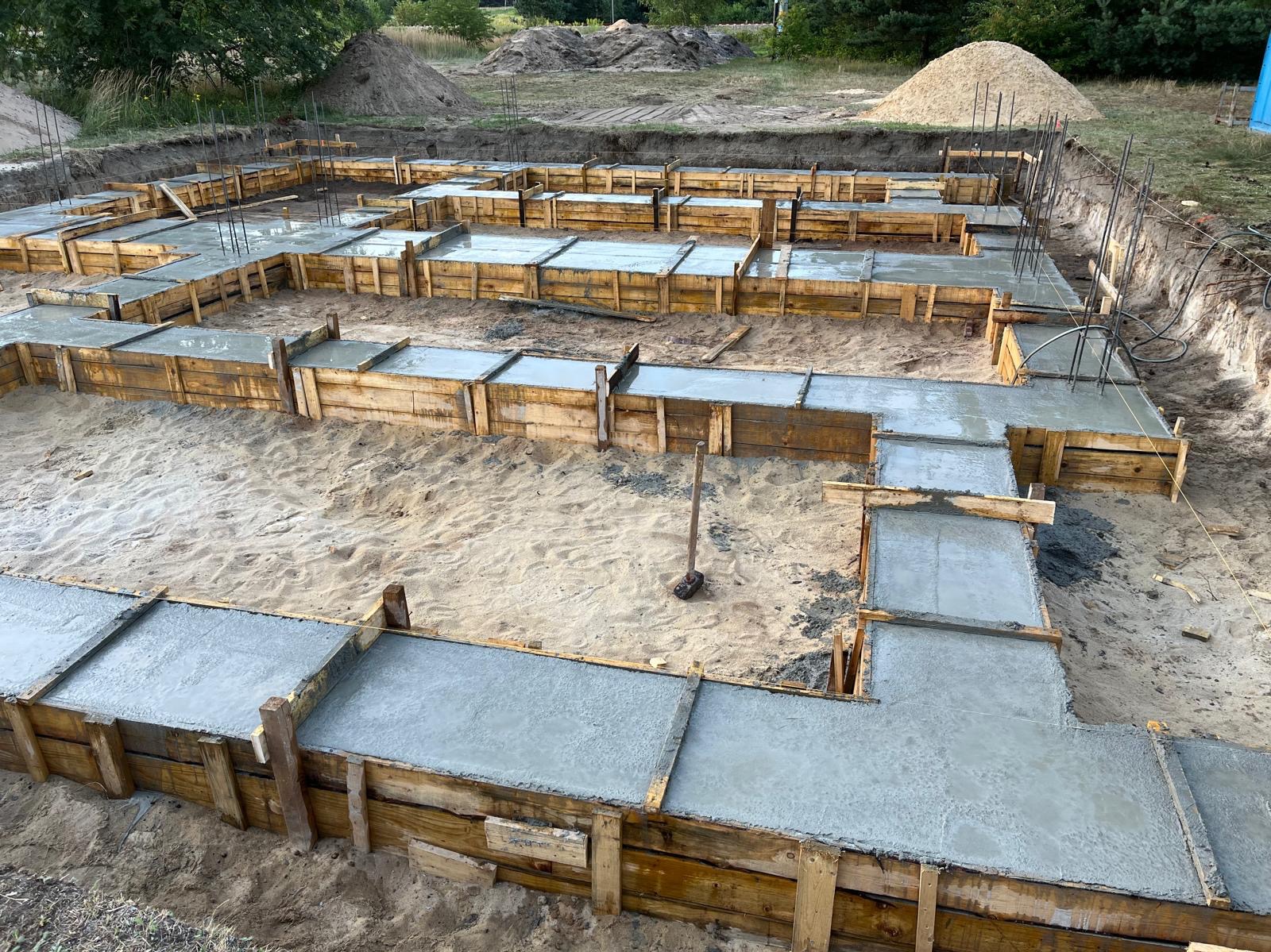 Wylany beton w drewnianym szalunku pod fundament budynku, widoczne wystające pręty zbrojeniowe i wbity młotek ciesielski w piasek.