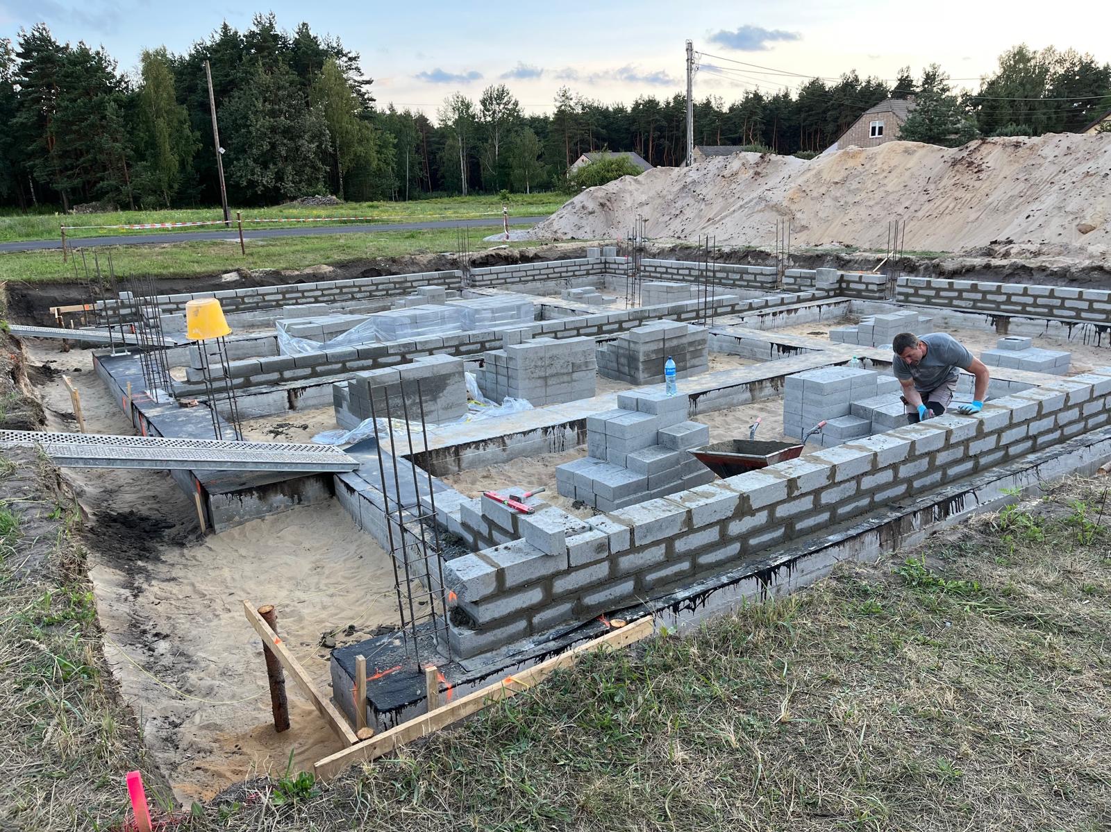 Budowa fundamentów z bloczków betonowych, widoczny murarz w trakcie pracy, teren budowy z piaskiem i drzewami w tle.