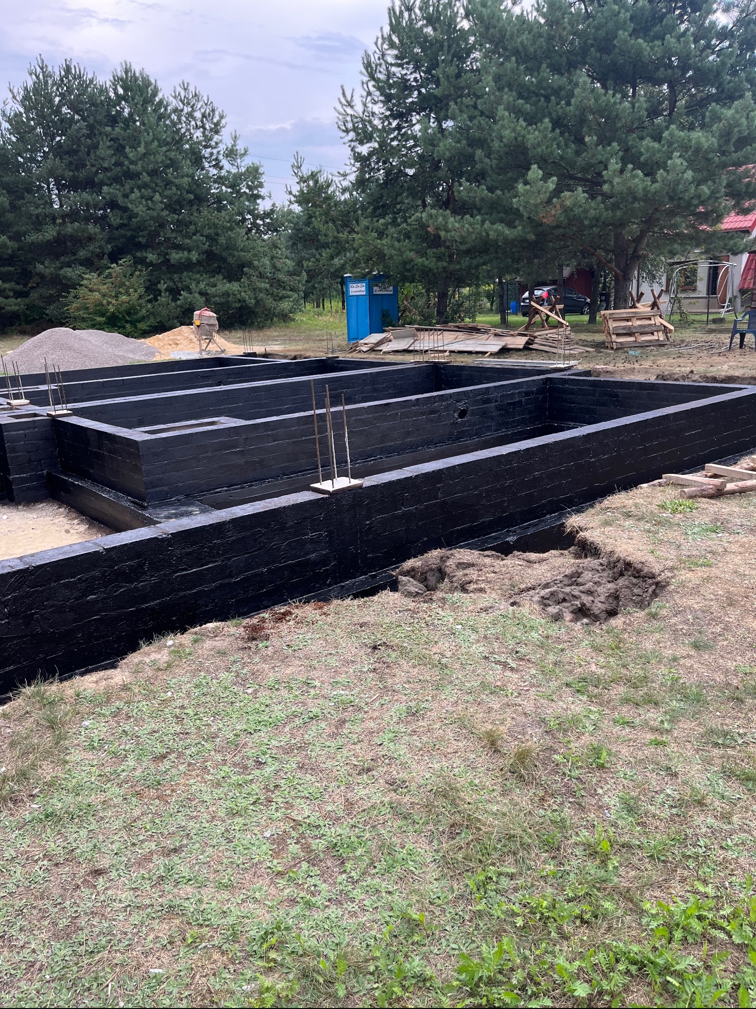 Zalane, czarną izolacją przeciwwilgociową, ściany fundamentowe z wystającymi prętami zbrojeniowymi na placu budowy, w tle widoczna betoniarka, przenośna toaleta i sterta piasku.