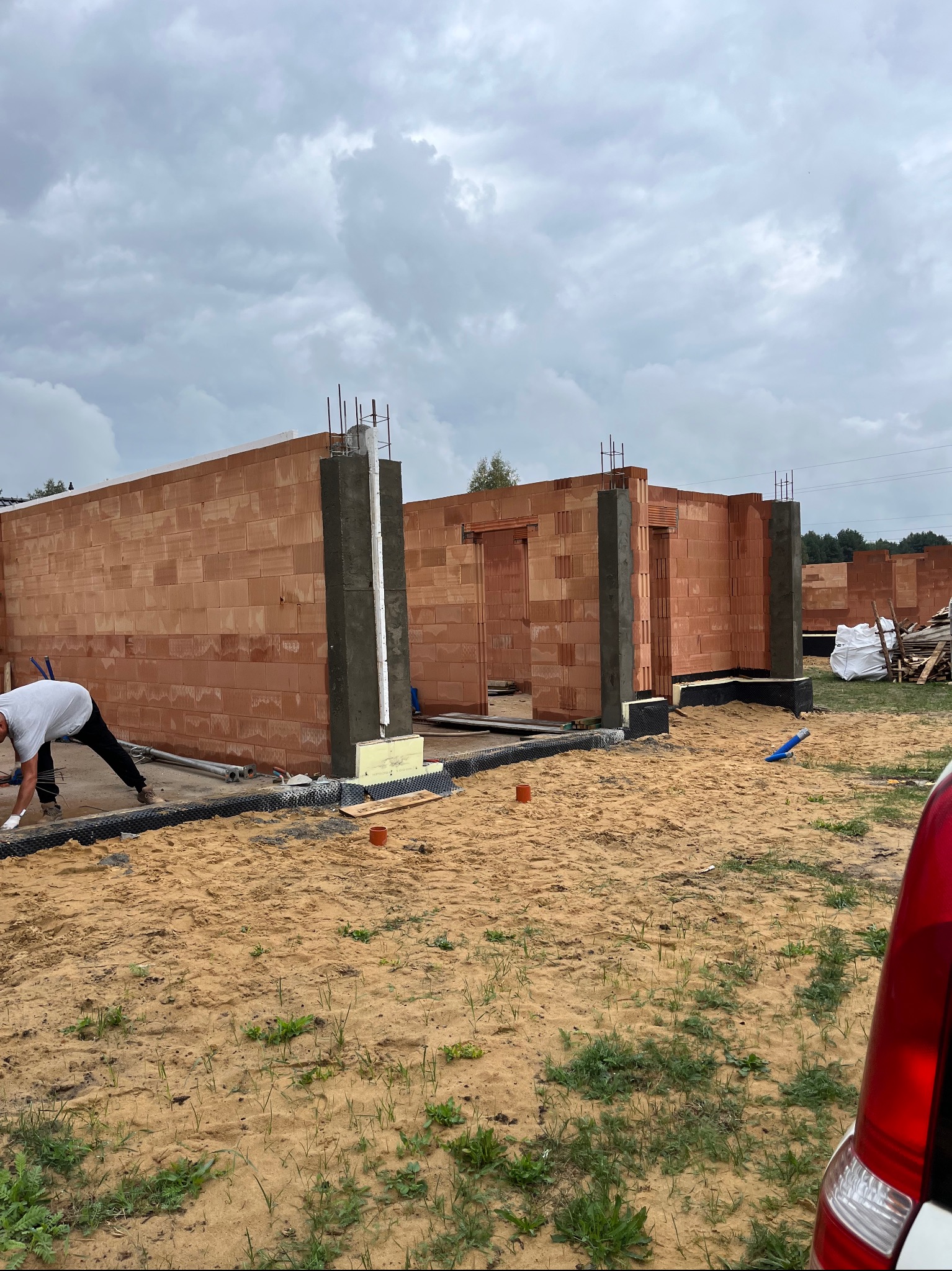 Budowa domu w trakcie: ściany z czerwonej cegły, fundamenty z izolacją, pracownik w białej koszulce w trakcie pracy, widoczne zbrojenia słupów, piaszczyste podłoże.
