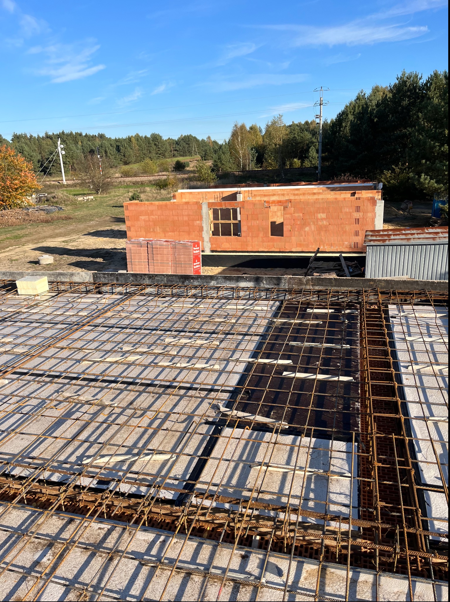 Widok z góry na zbrojenie stropu i fragment wznoszonej ściany z czerwonej cegły ceramicznej Porotherm, z widocznymi otworami okiennymi, na tle zieleni drzew i błękitnego nieba.