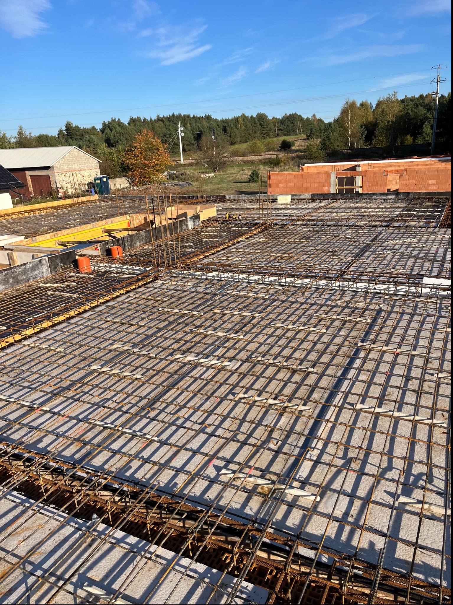 Zbrojenie płyty fundamentowej domu jednorodzinnego, widok z góry na siatkę z prętów stalowych, szalunki drewniane w tle, błękitne niebo.