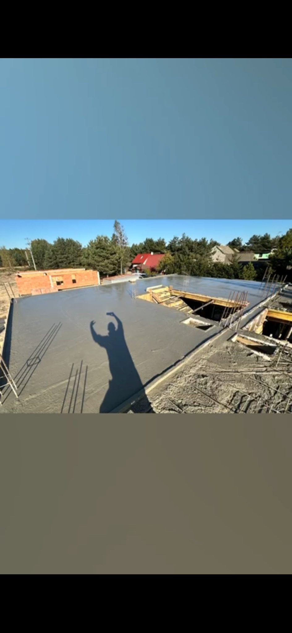 Świeżo wylany, gładki betonowy strop z widocznym zbrojeniem i deskowaniem, cień osoby z uniesionymi rękami na powierzchni, w tle zabudowa jednorodzinna i zieleń.