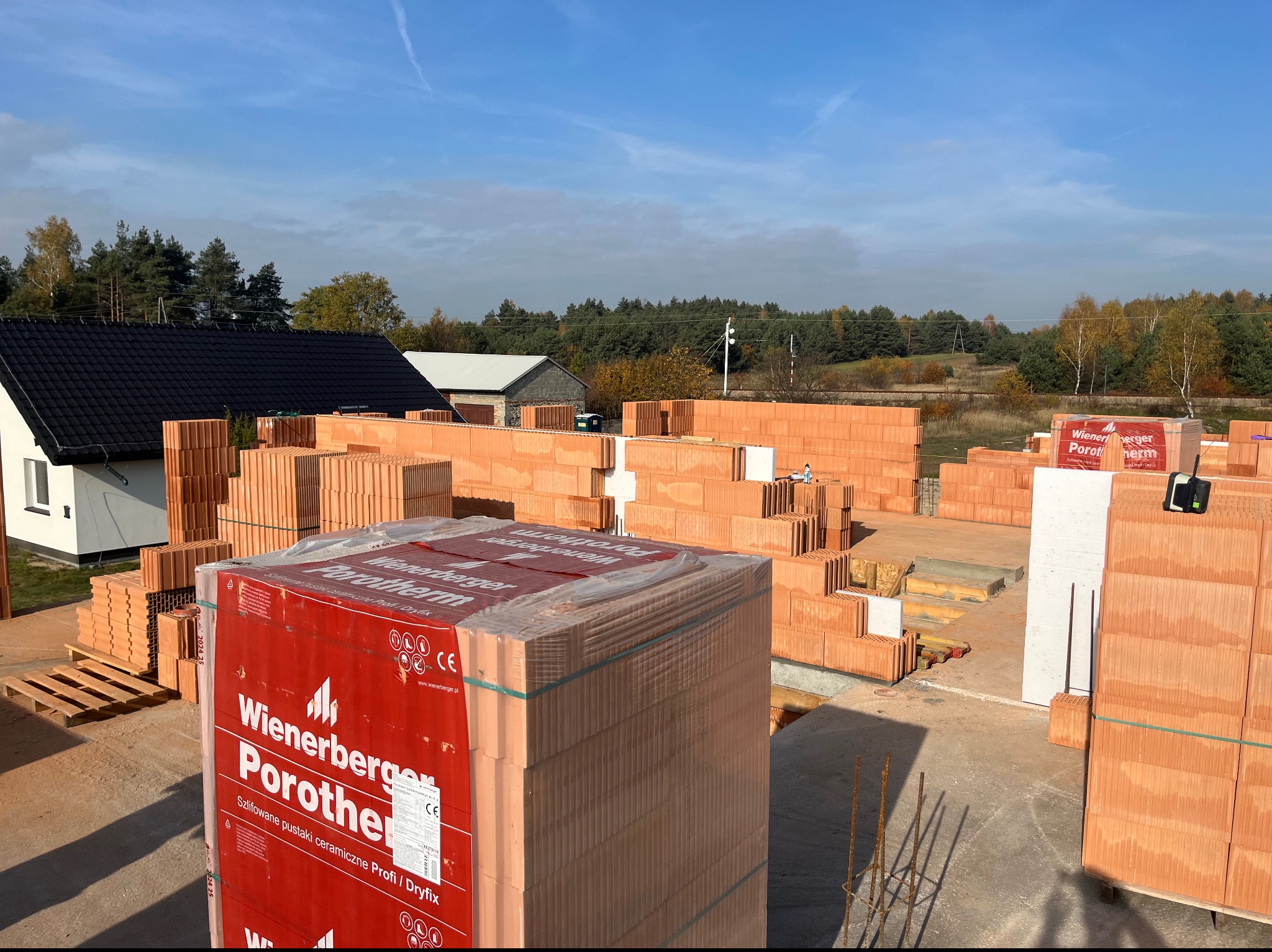 Budowa domu z pustaków ceramicznych Porotherm Wienerberger, widok na częściowo wymurowane ściany, palety z materiałem budowlanym i otaczający krajobraz w słoneczny dzień.