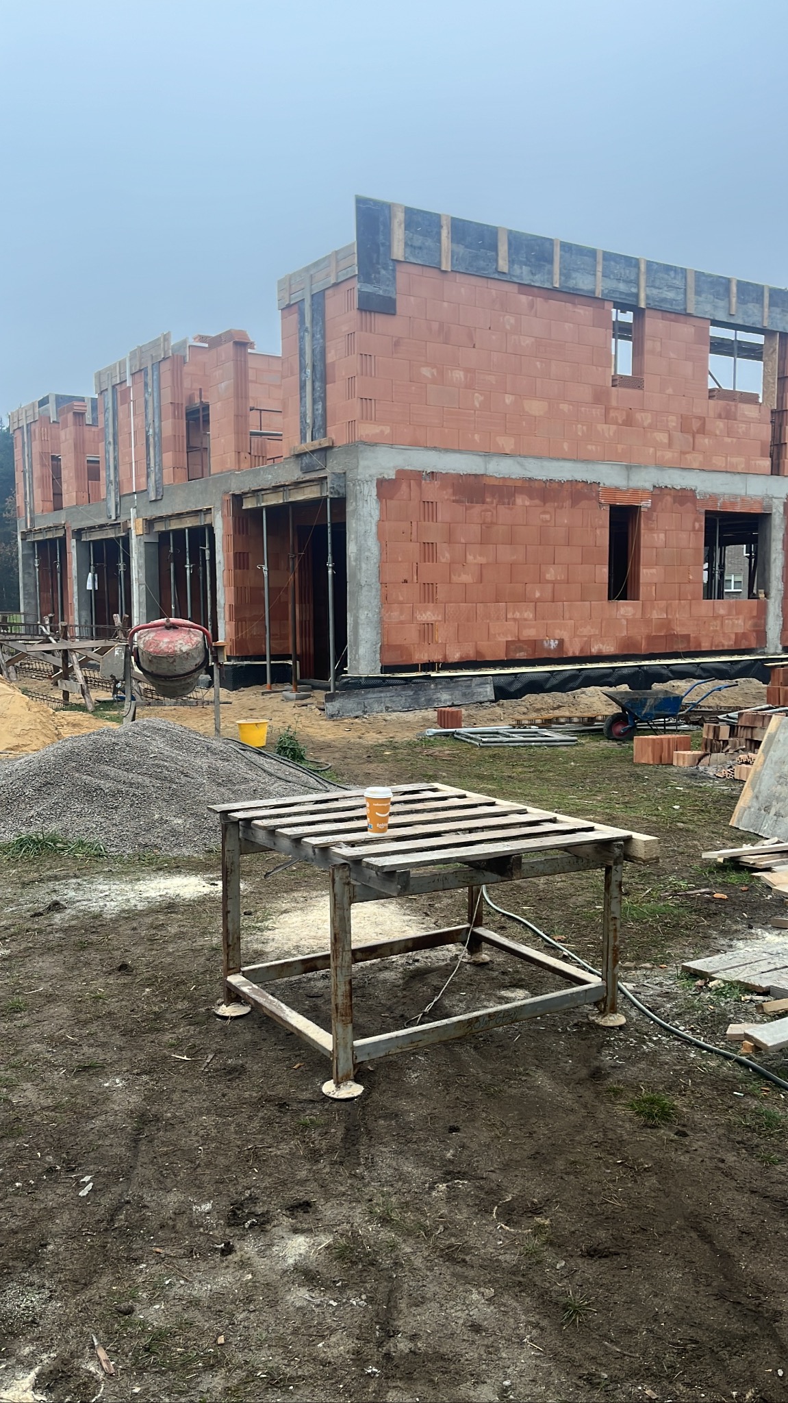 Stan surowy zamknięty szeregu domów z czerwonej cegły, widok z zewnątrz, kubek z kawą na metalowym stole na pierwszym planie, mieszarka do betonu i hałda żwiru w tle.