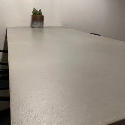 KRIS-STUC Krystian Maczukin - Blat stołu z szarego betonu architektonicznego z delikatną fakturą, na którym stoi mały kaktus w doniczce z efektem ombre, na tle białej ściany.