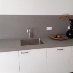 KRIS-STUC Krystian Maczukin - Szary blat kuchenny z minimalistycznym zlewem, białe szafki z podłużnymi uchwytami, dekoracyjna czarna waza i drewniana misa z orzechami, szara ściana nad blatem, gniazdka elektryczne.