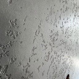 KRIS-STUC Krystian Maczukin - Szara ściana z dekoracyjną fakturą, widoczny otwór z przewodami elektrycznymi w dolnym rogu.