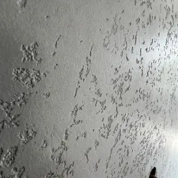 Szara ściana z dekoracyjną fakturą, widoczny otwór z przewodami elektrycznymi w dolnym rogu.