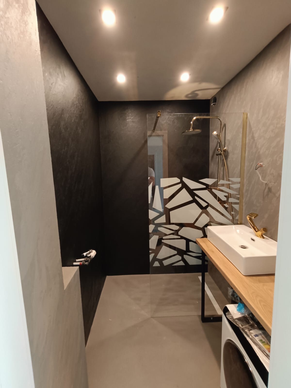Nowoczesna łazienka z czarno-szarymi ścianami, kabiną prysznicową z geometrycznym wzorem i złotą armaturą, umywalka nablatowa na drewnianym blacie.