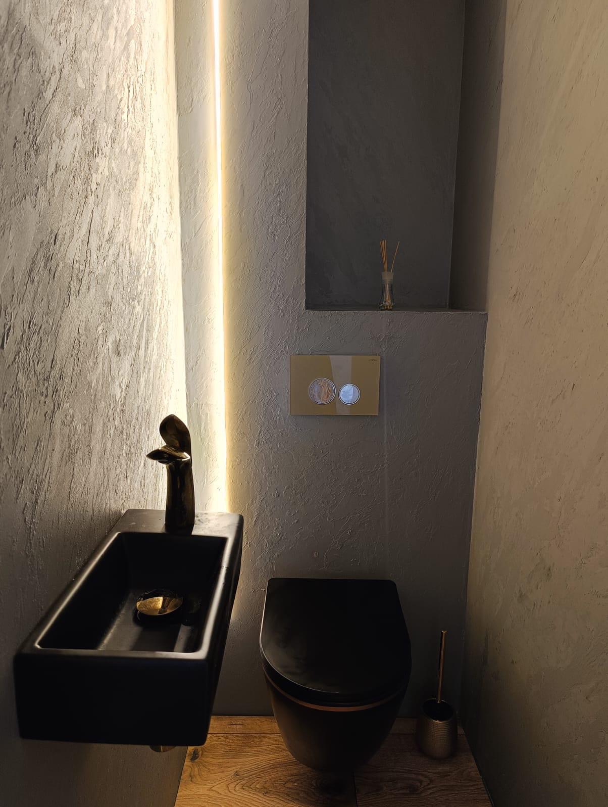 Nowoczesna łazienka z czarną umywalką nablatową, złotą baterią w kształcie ptaka i wiszącą toaletą w kolorze czarnym, oświetlona pionową lampą LED na tle ściany wykończonej mikrocementem.